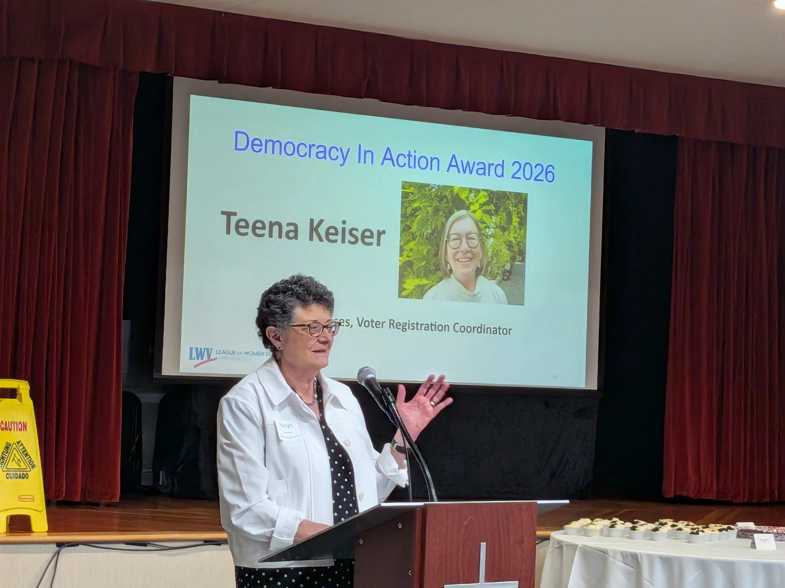 A woman speaking at a podium during an award ceremony titled 'Democracy In Action Award 2026'. There is a large screen behind her displaying her name, Teena Keiser, and her photo. The room has red curtains and caution signs on the side.