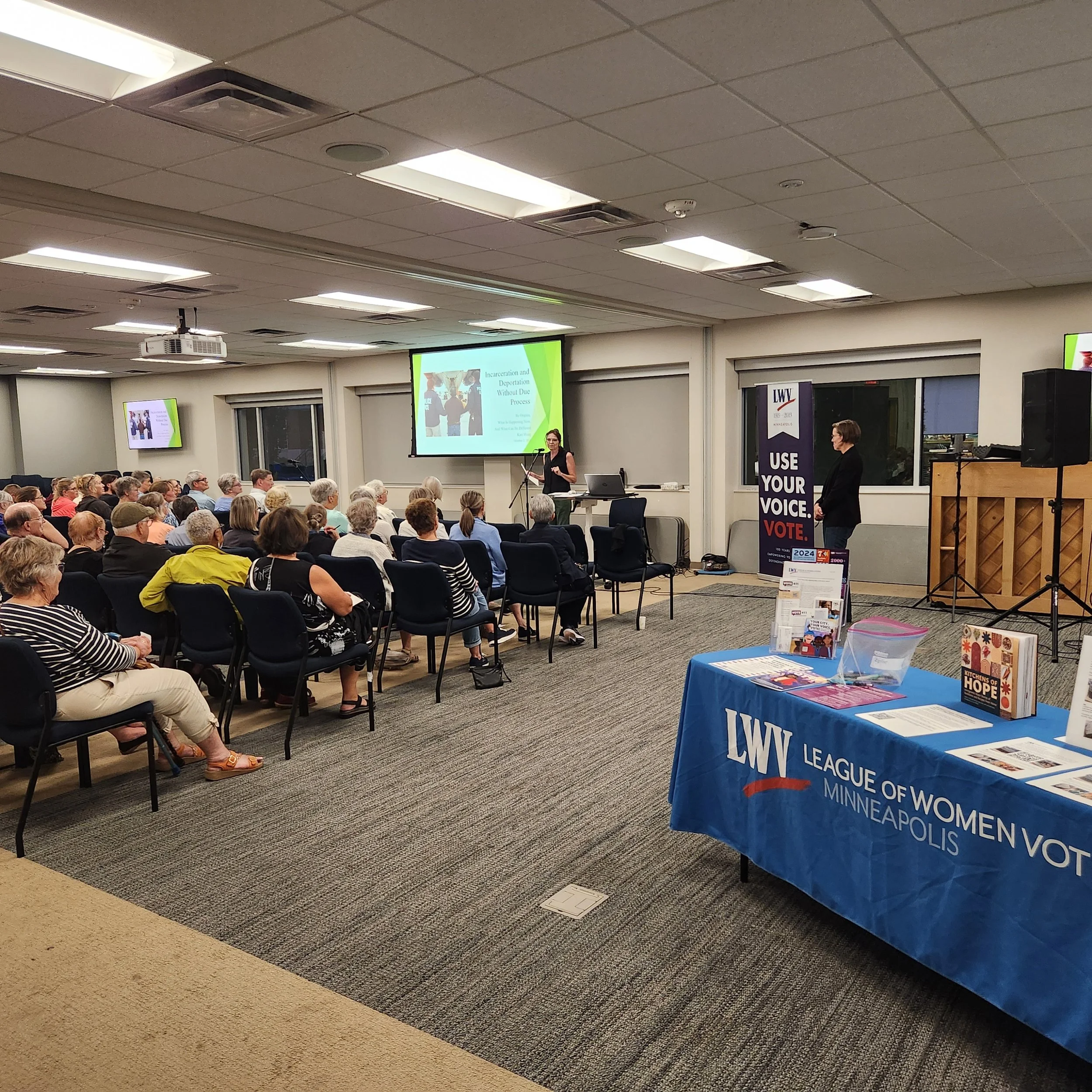 View of a conference room with a large crowd of people sitting and a woman speaking at a podium. There are multiple screens displaying a presentation and a table with promotional materials and banners, including one with the text 'Leauge of Women Voters of Minneapolis, VOTE'.