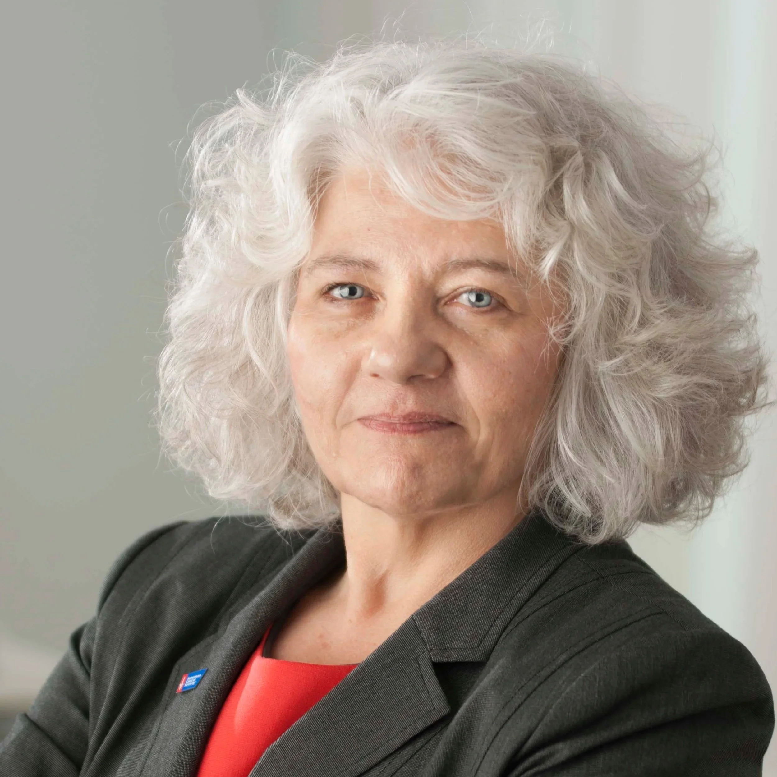 Close-up of an elderly woman with curly white hair, light skin, and blue eyes, wearing a dark blazer and a red top, against a plain background.