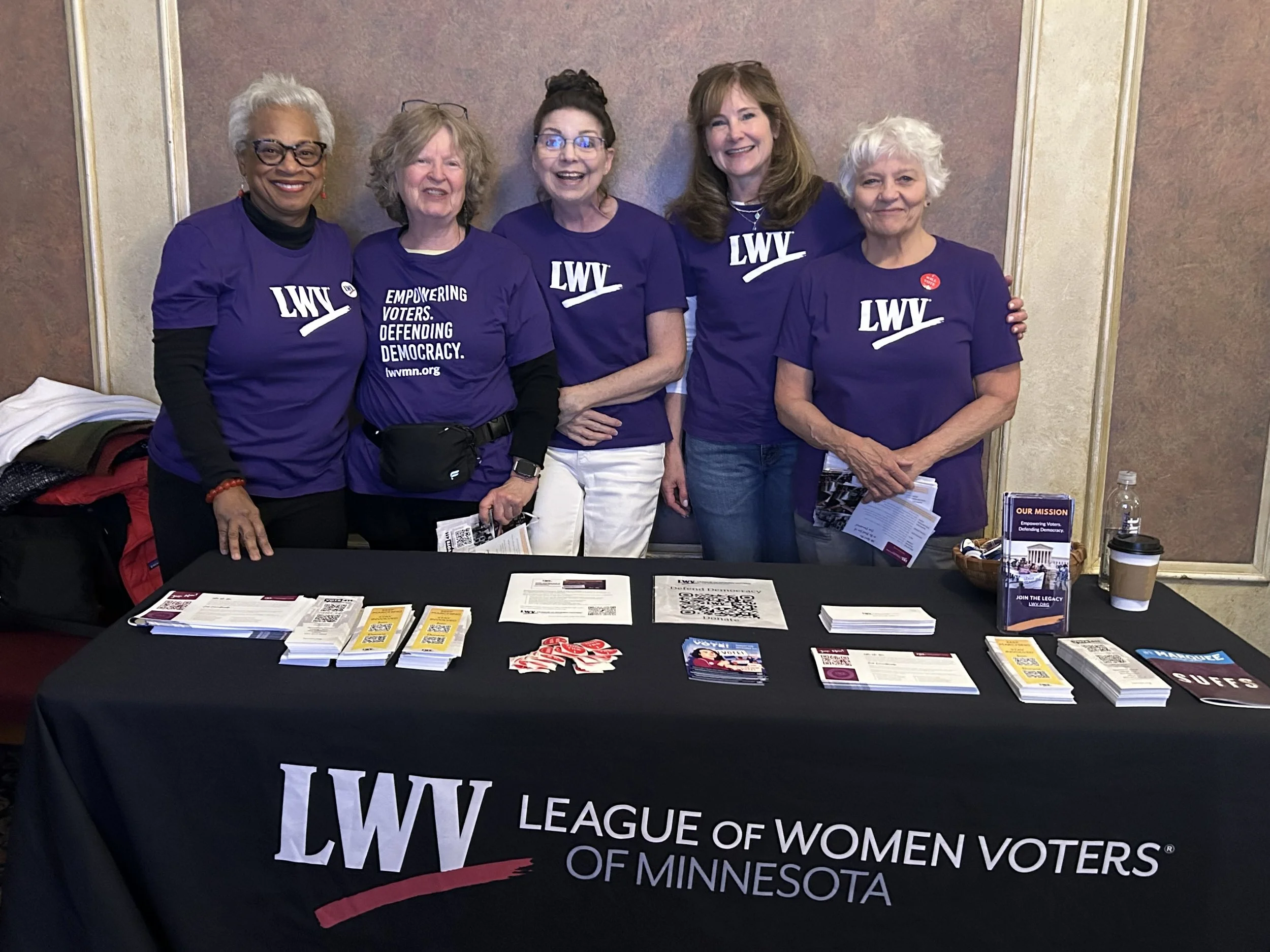 Five women standing behind a table at a League of Women Voters event, all wearing purple shirts with the LWV logo and slogans supporting voting rights. The table has brochures, pamphlets, and campaign materials.