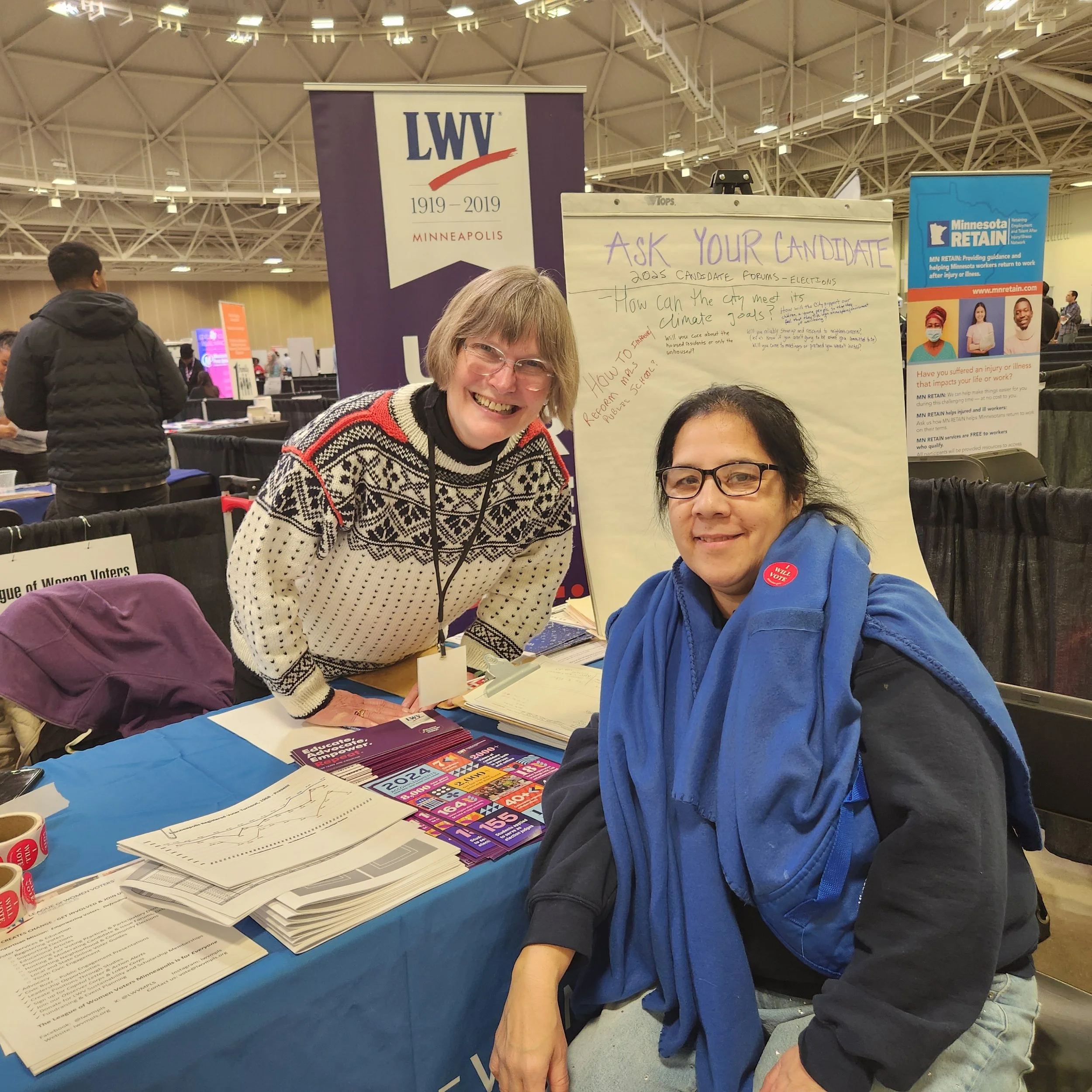 Two smiling women at a table with political campaign materials, with signs about voting and voting questions behind them at an indoor event.