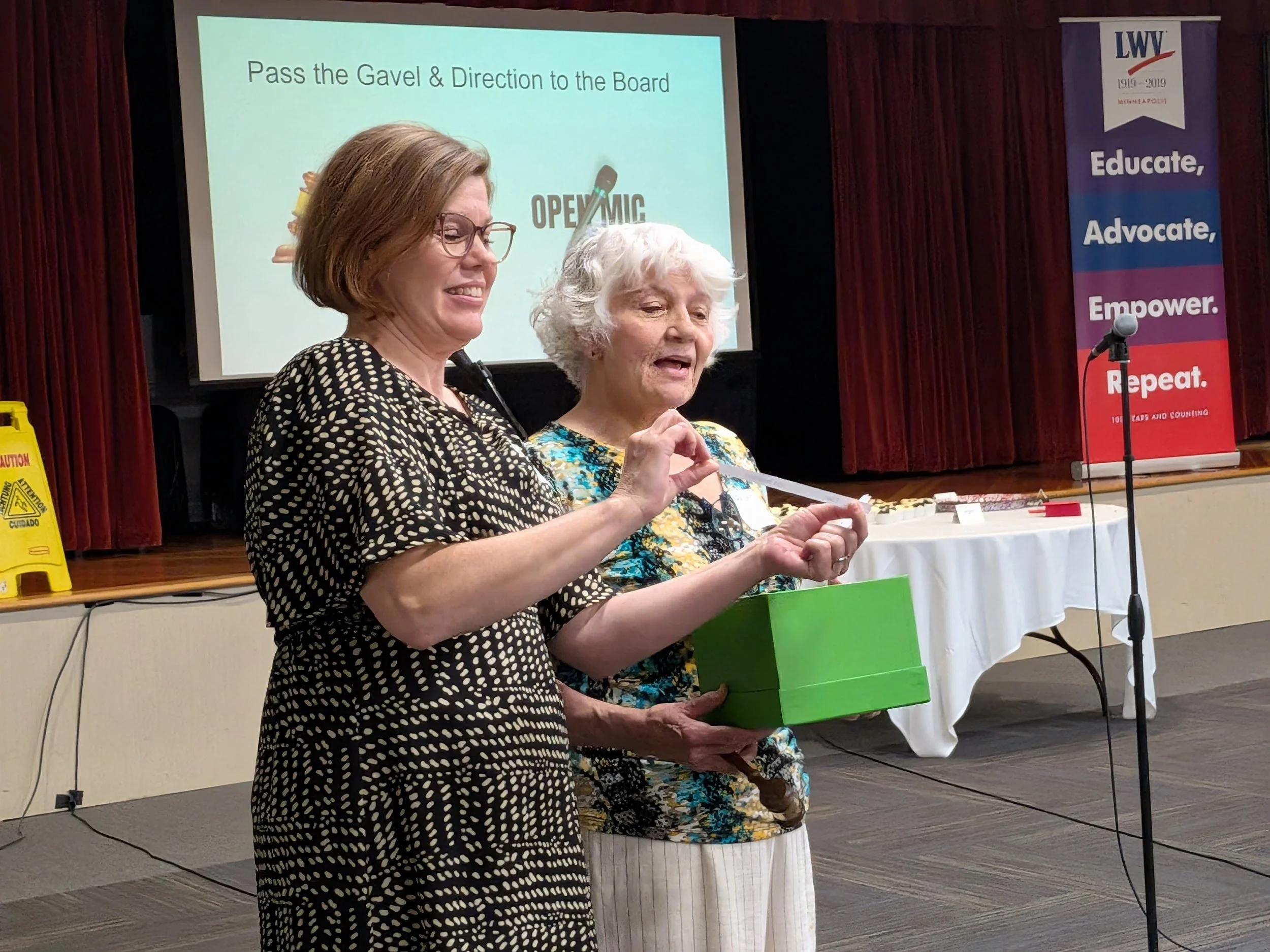 Two women standing in front of a presentation screen, one holding a green box, with a banner that reads 'LWV' and 'Educate, Advocate, Empower, Repeat'.