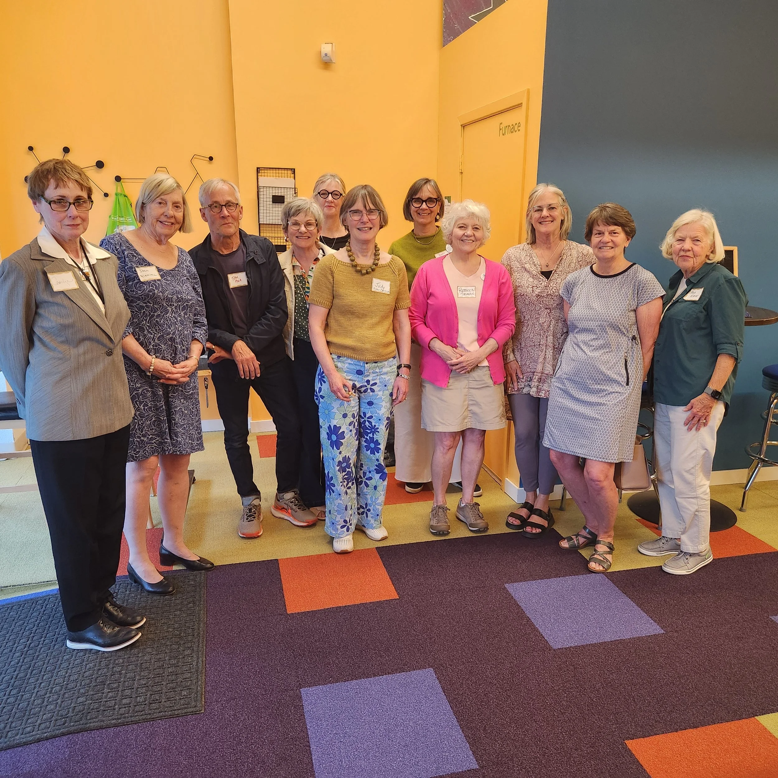 A group of people gathered indoors in front of colorful walls, some wearing glasses and smiling, with various name tags visible, standing on a multicolored carpet.