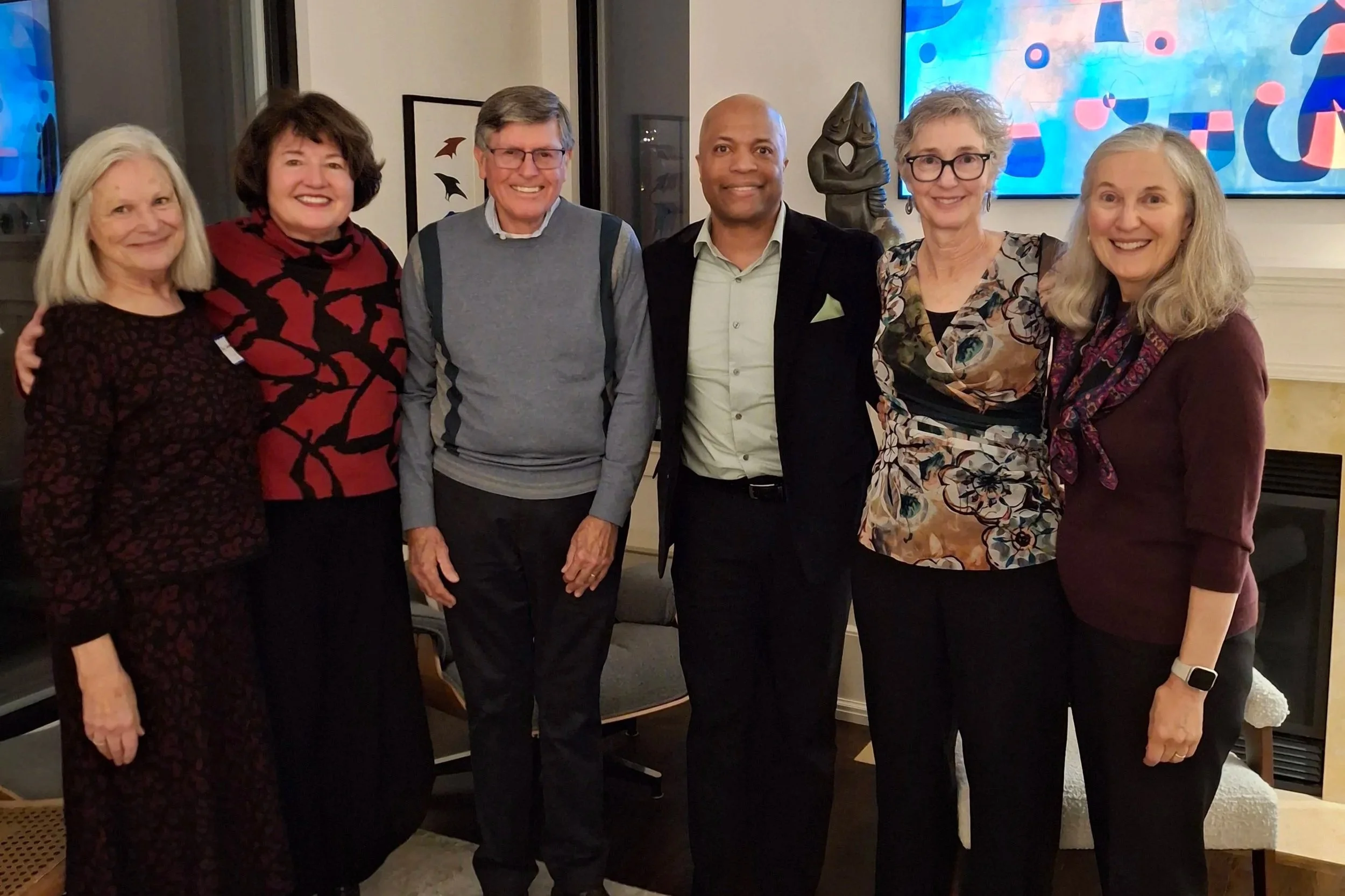 Group of seven people standing together indoors, smiling at the camera, with a colorful abstract painting on the wall behind them.