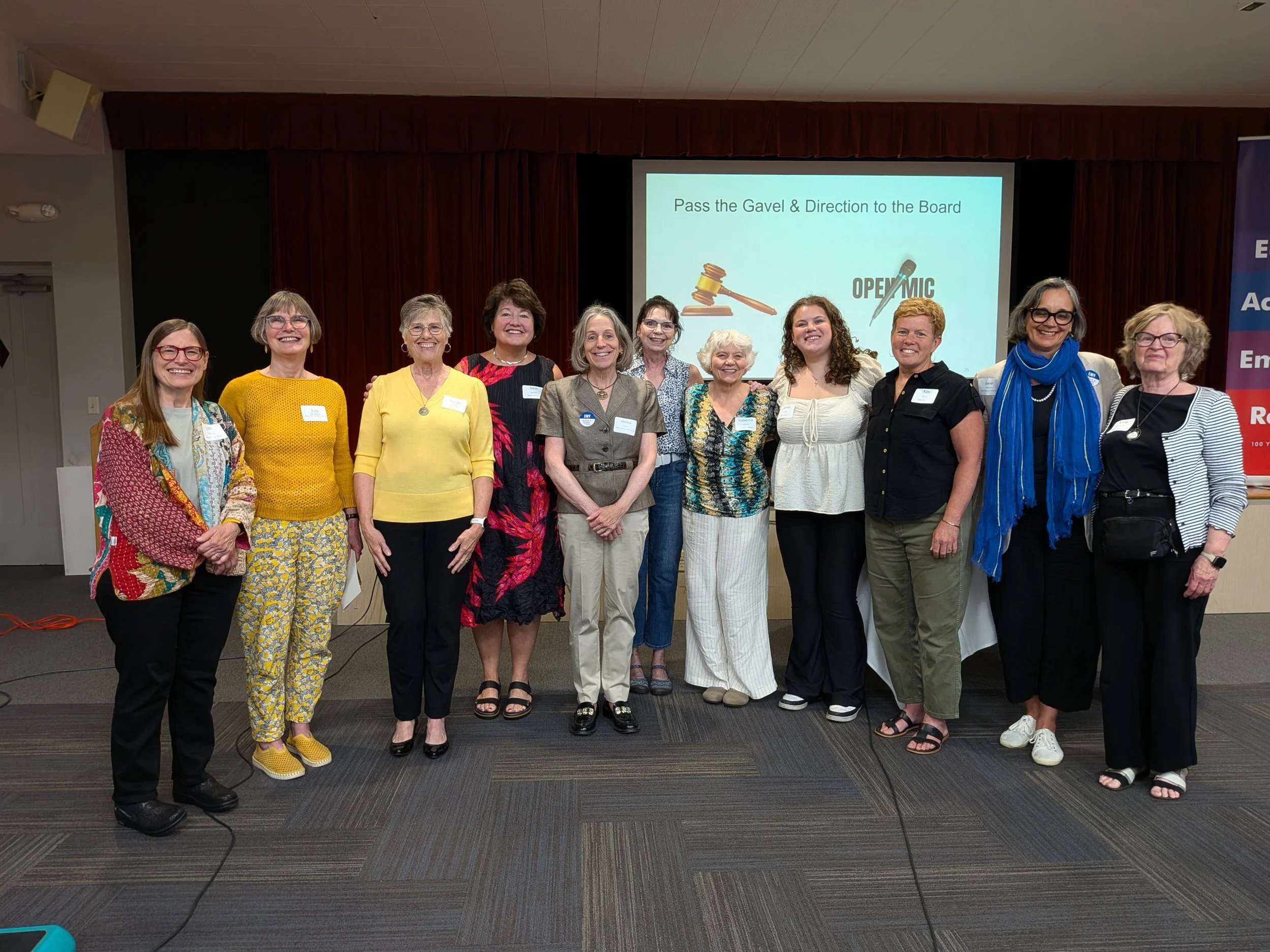 A diverse group of fifteen women standing together on a stage in front of a large screen displaying the text "Pass the Gavel & Direction to the Board" at what appears to be a professional conference or meeting.