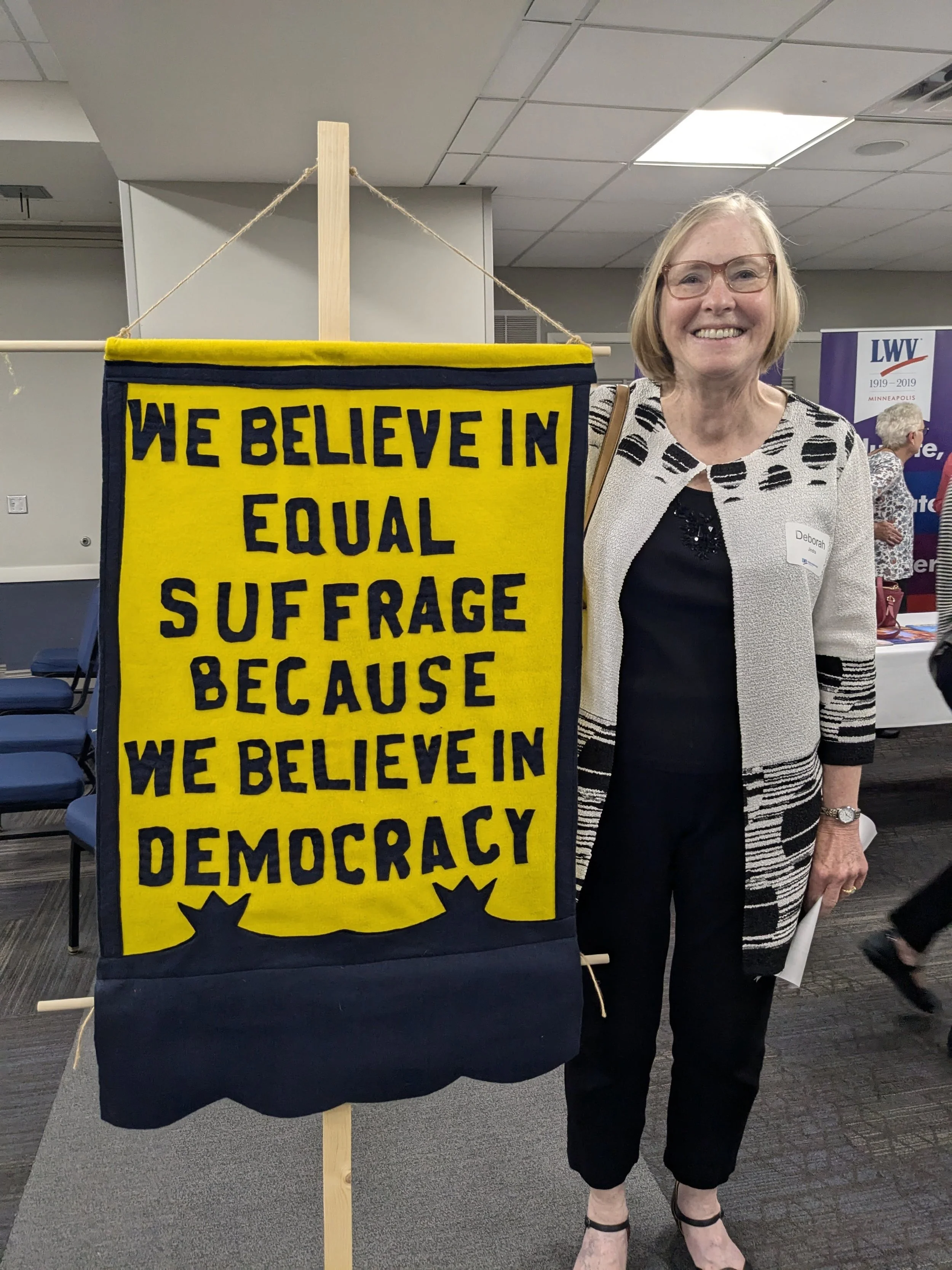 A woman with short blonde hair and glasses smiling, standing next to a yellow and black sign that reads 'We believe in equal suffrage because we believe in democracy'. She is wearing a black dress with a white and black patterned blazer and black heels. The setting appears to be indoors, possibly at a conference or event, with other people and banners in the background.