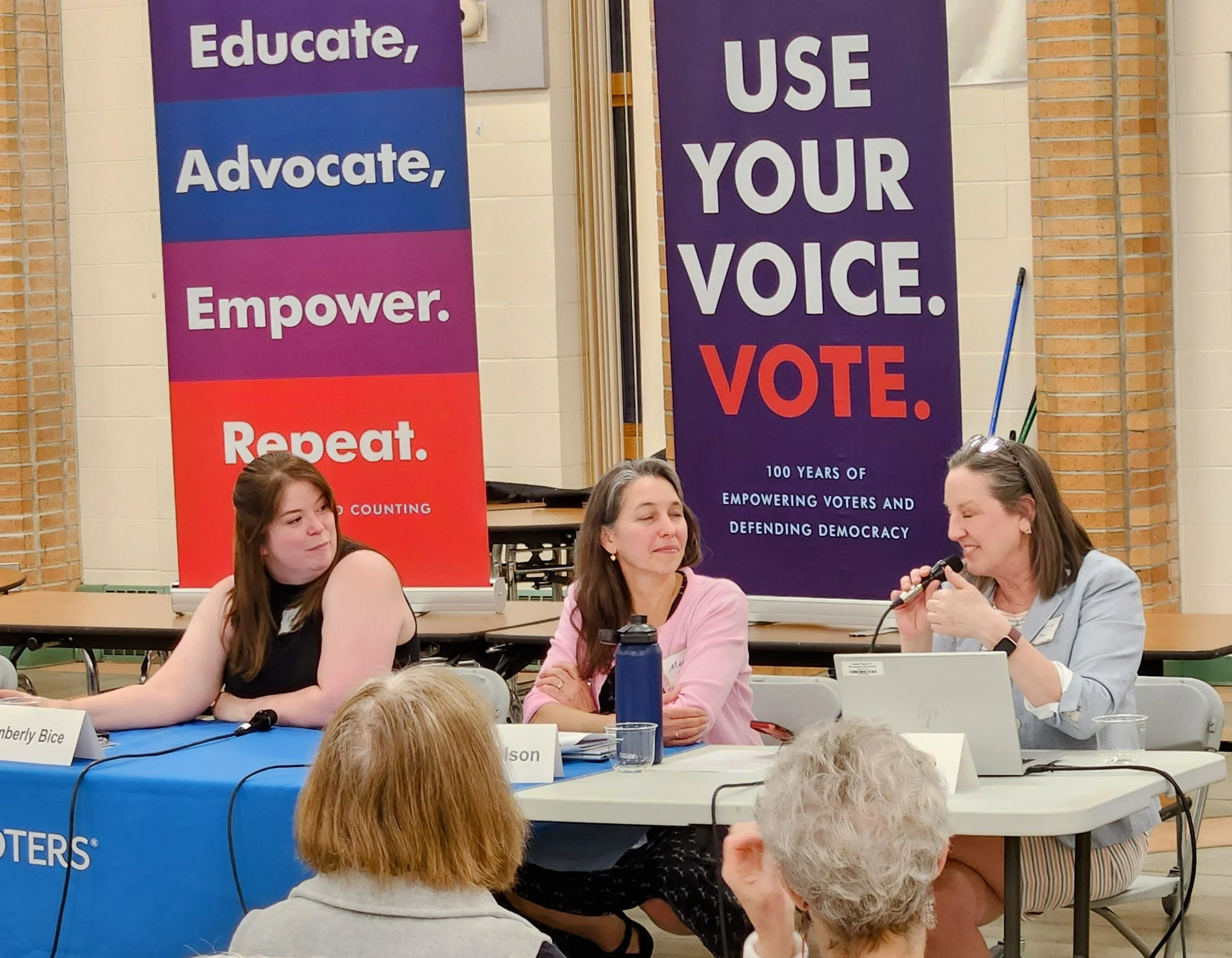 Group of women sitting at a table during a panel discussion or meeting, with large banners behind them displaying messages about educating, advocating, empowering, and voting.