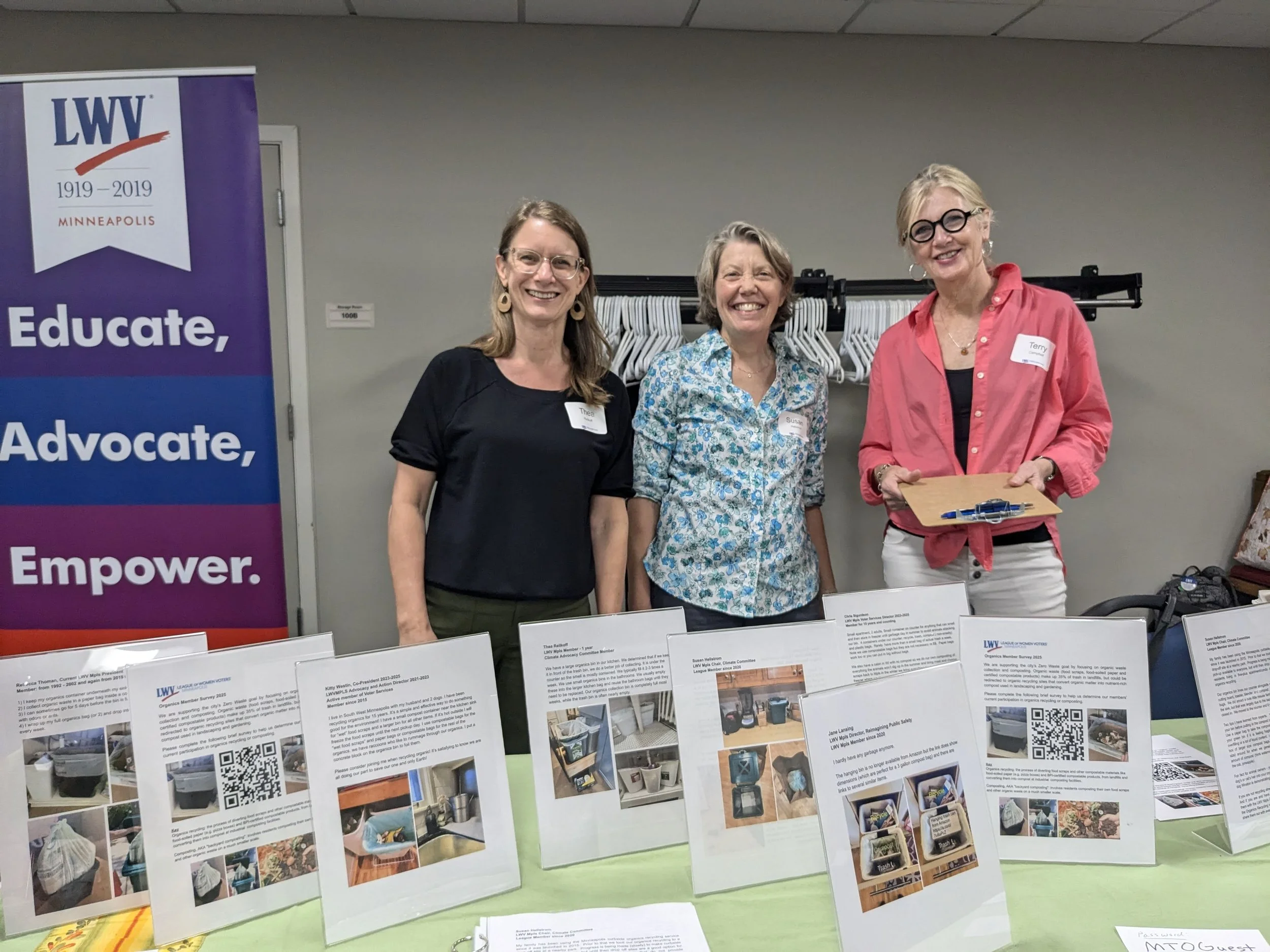 Three women standing in front of a display board and a purple banner that reads 'LWV 1919-2019 Minneapolis'. All women are smiling and wearing name tags. The display board contains various printed materials and photographs, with a coat rack and some 