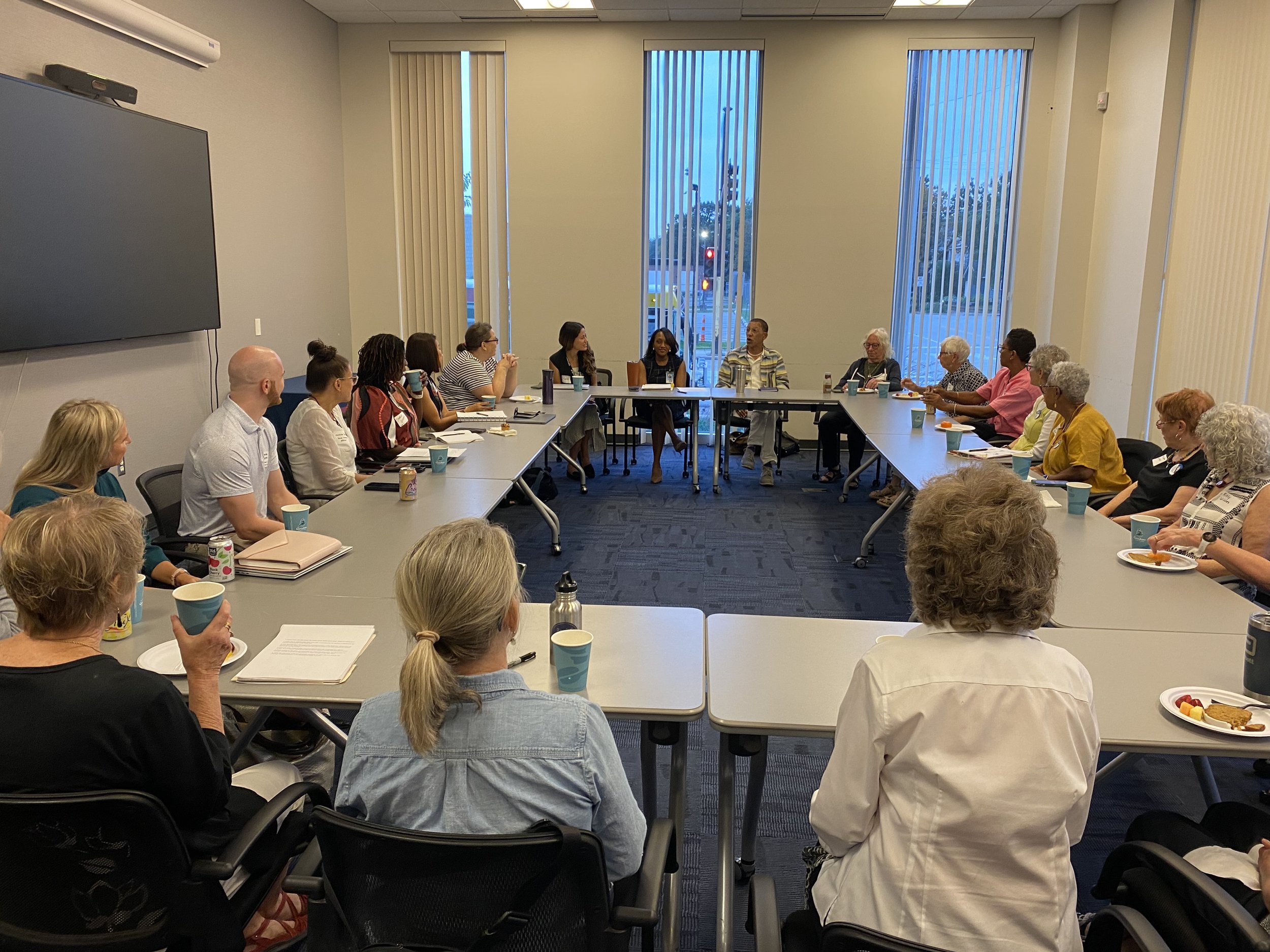 A diverse group of approximately 16 people, mostly women, seated around a large U-shaped conference table in a brightly lit room with large windows and vertical blinds, engaging in a discussion.