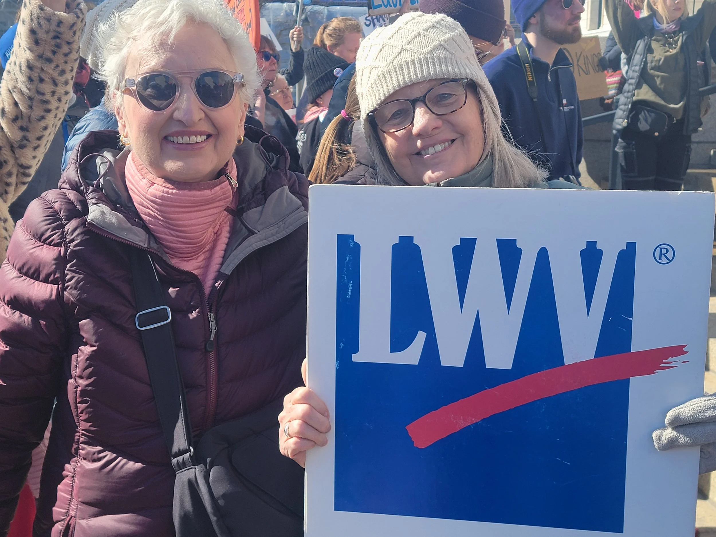 Two women at a protest or rally, smiling and holding a sign that partially reads 'LIV' with a red, white, and blue design, surrounded by other protesters holding signs and banners.
