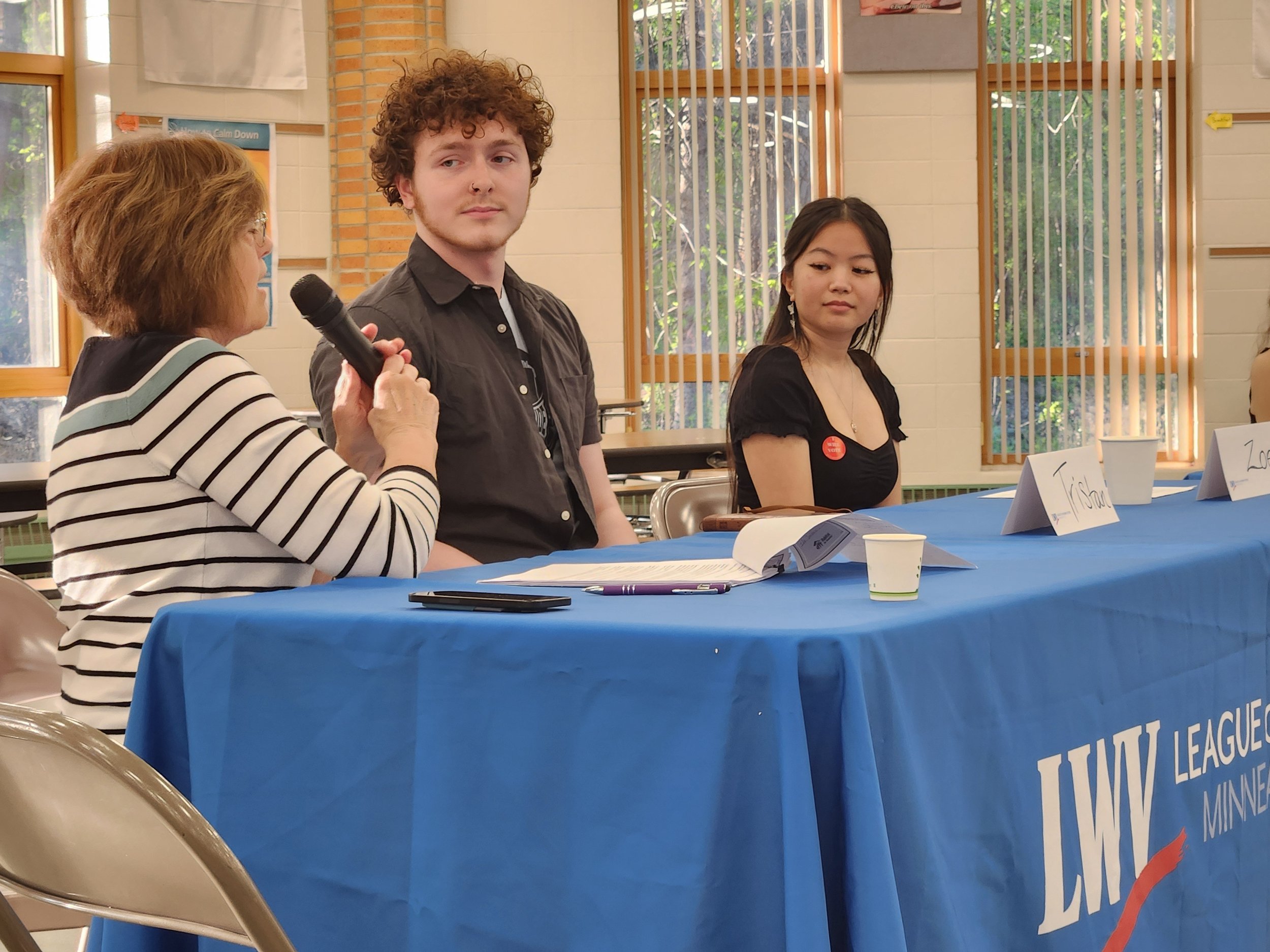 A woman holding a microphone is speaking at a table with two young women. The young woman on the left has curly hair and is looking toward the woman, while the one on the right has straight dark hair and is wearing earrings and a badge. There are nam