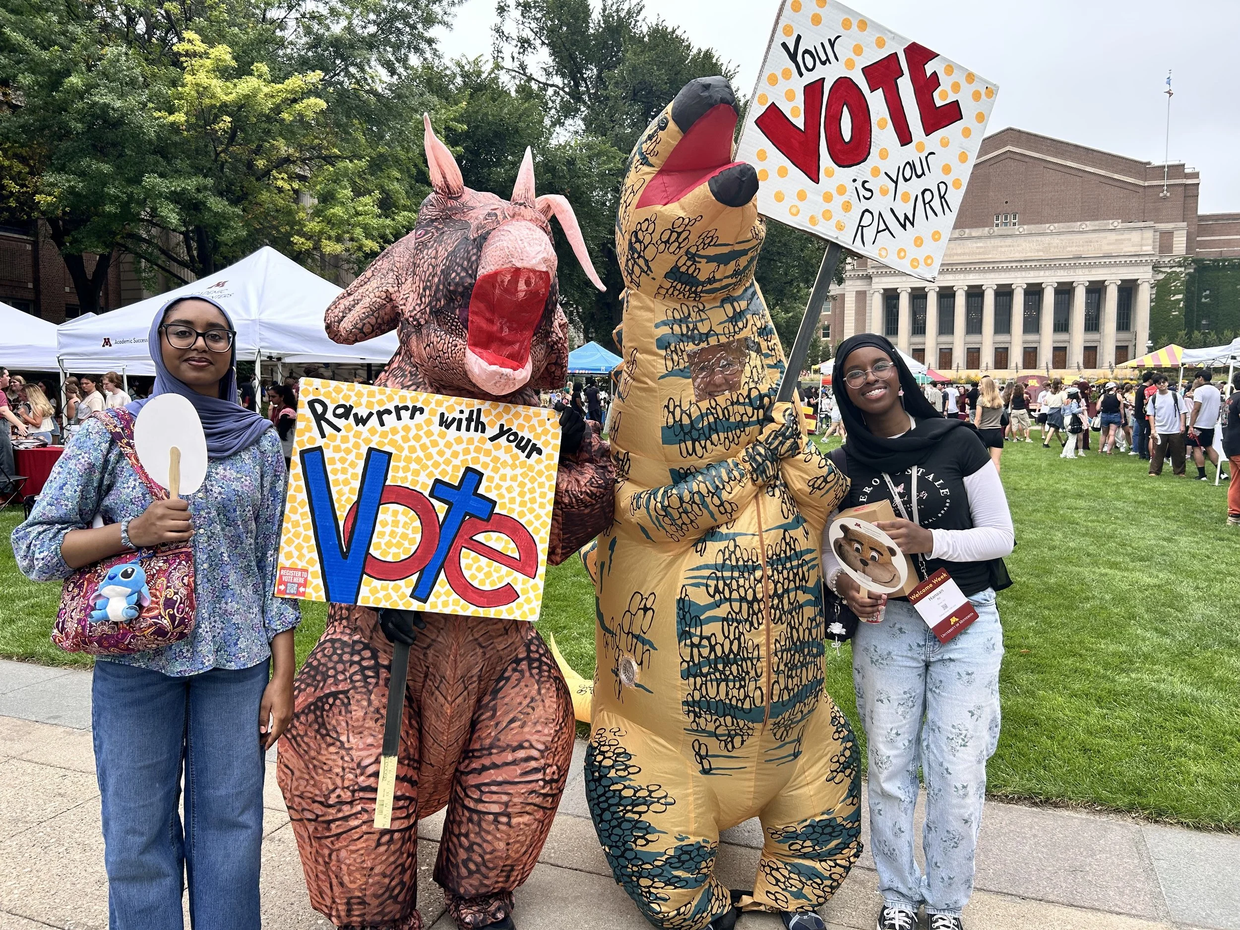 Two women and two people in inflatable dinosaur costumes holding signs at an outdoor event. One sign says 'Your vote is your RAWRR,' and the other says 'Rawrr with your VOTE.' There are tents and many people in the background.