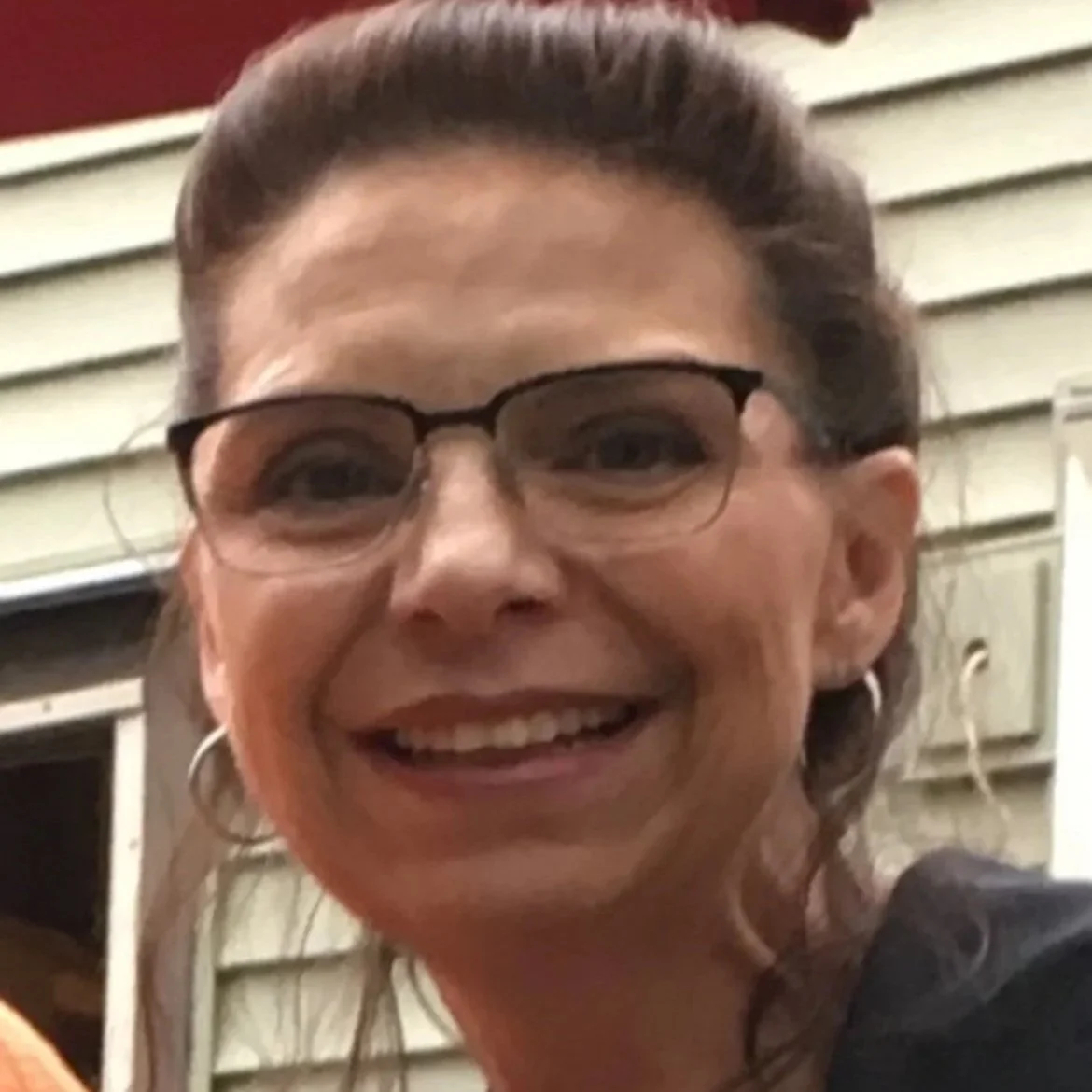 A woman smiling, wearing glasses and hoop earrings, in front of a house with siding.