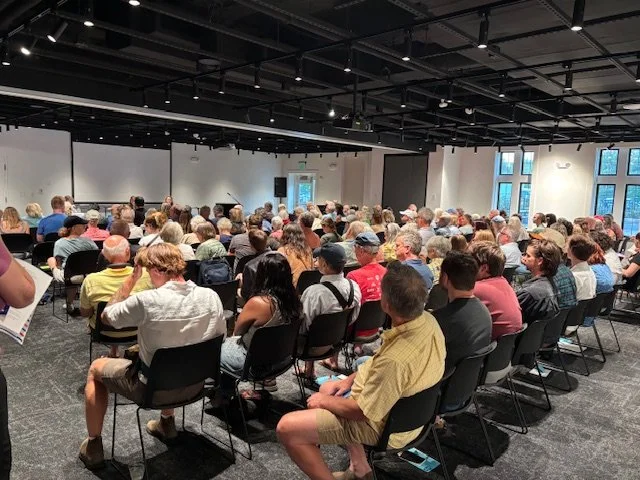 A large group of people attending a conference or seminar in a well-lit room with large windows.