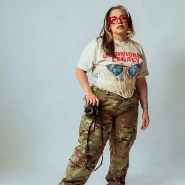 Young woman with long brown hair, wearing red glasses, a t-shirt with red text, camouflage pants, and a corset, holding a camera, standing against a plain white background.