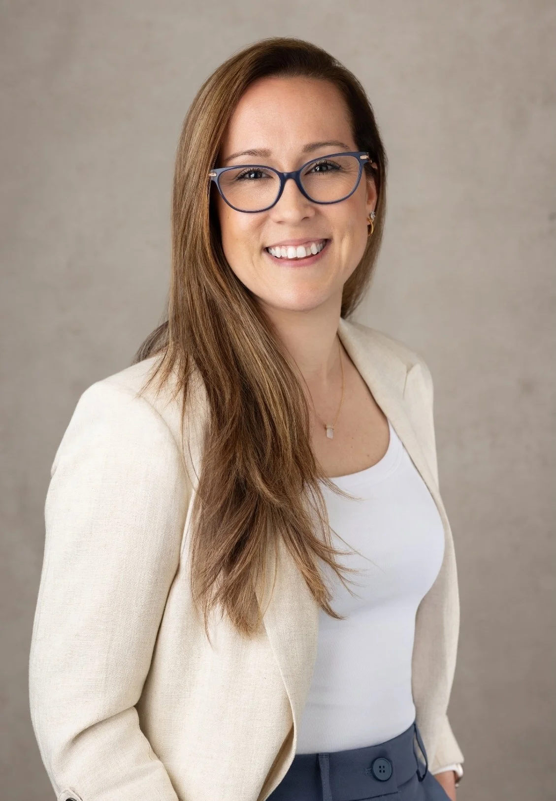 Dr. Kristina Burke, therapist based in Garden City, NY, with long brown hair, wearing glasses, a white top, beige blazer, and dark pants, smiling at the camera.