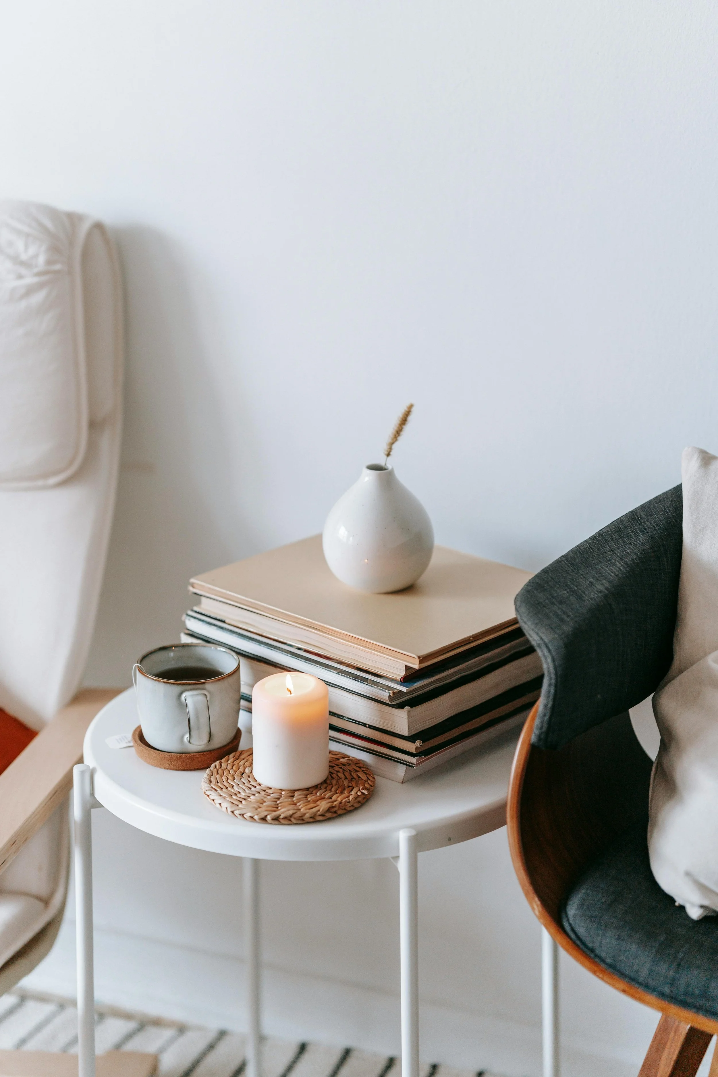 A side table with a white top, three stacked notebooks, a white vase with a dried flower, a mug of coffee on a cork coaster, a white candle, and a woven placemat. A gray and white chair with a wooden armrest is next to the table. Part of a beige sofa is visible on the left. The background features a plain white wall.