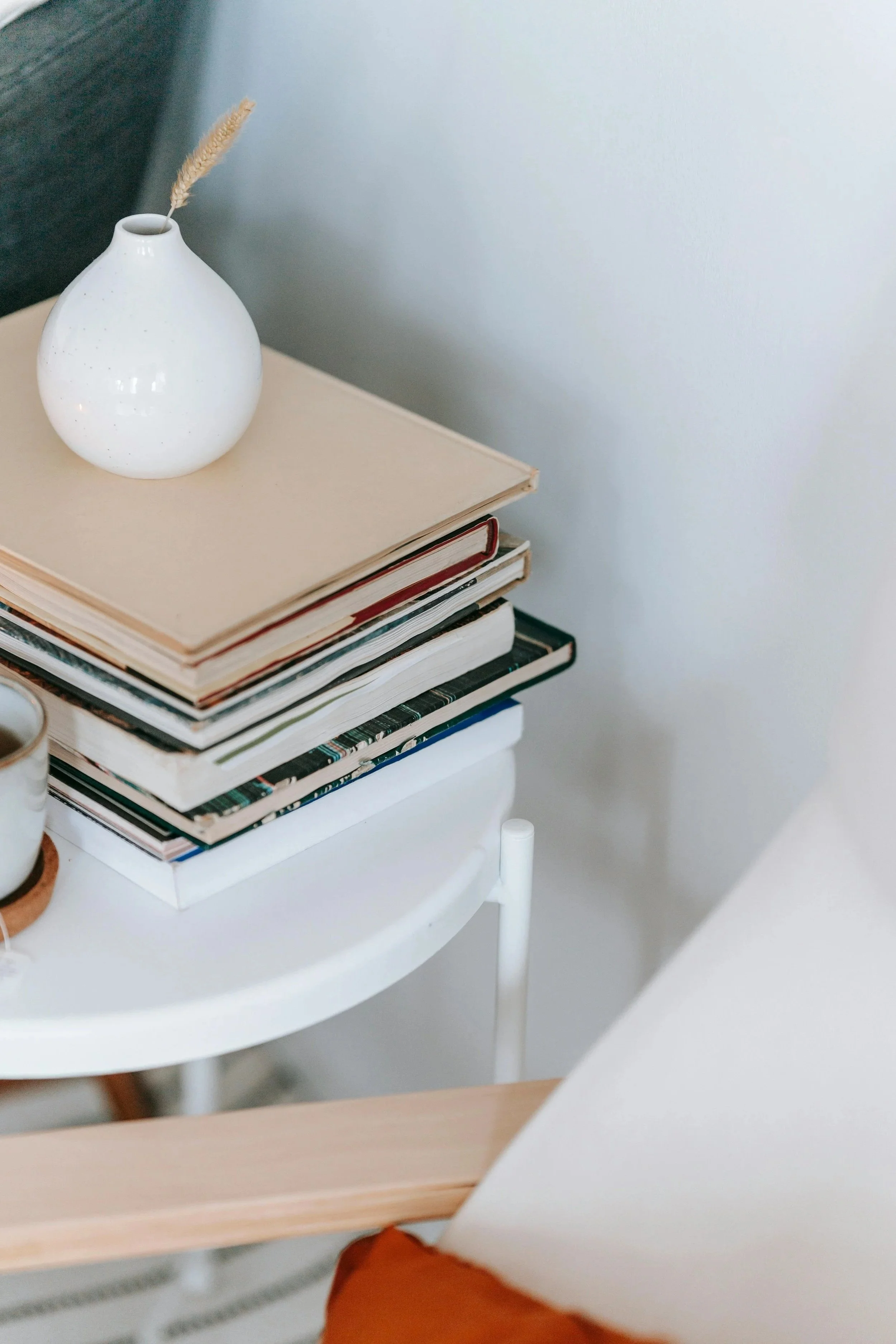 White vase with a single dried grass stalk on top of a stack of books on a white table.
