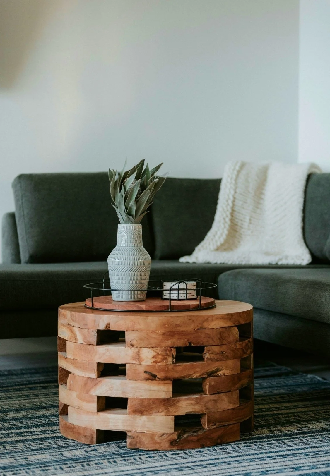 Living room with a dark gray sofa, a white knit blanket, a wooden coffee table with a vase holding green plants, and a tray with candles on the table. The floor has a patterned rug.