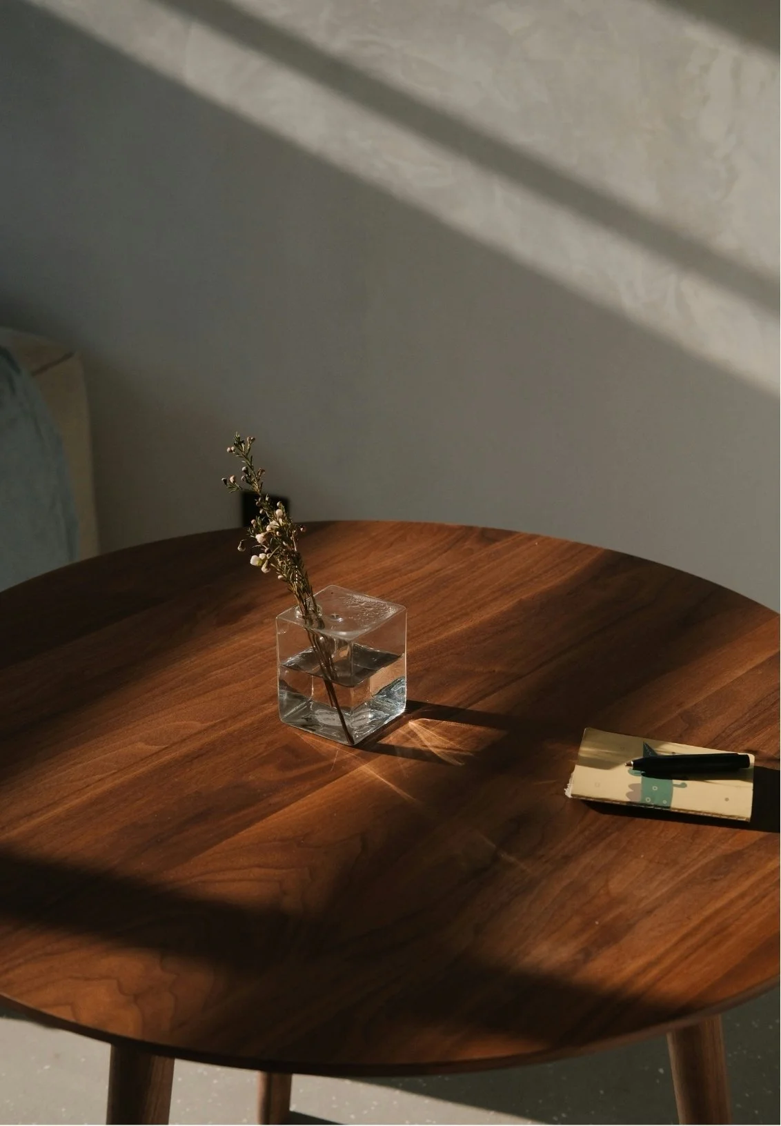 A wooden round table with a small square glass vase containing a sprig of dried flowers, opposite a closed notebook with a black pen on top, in natural light casting shadows.