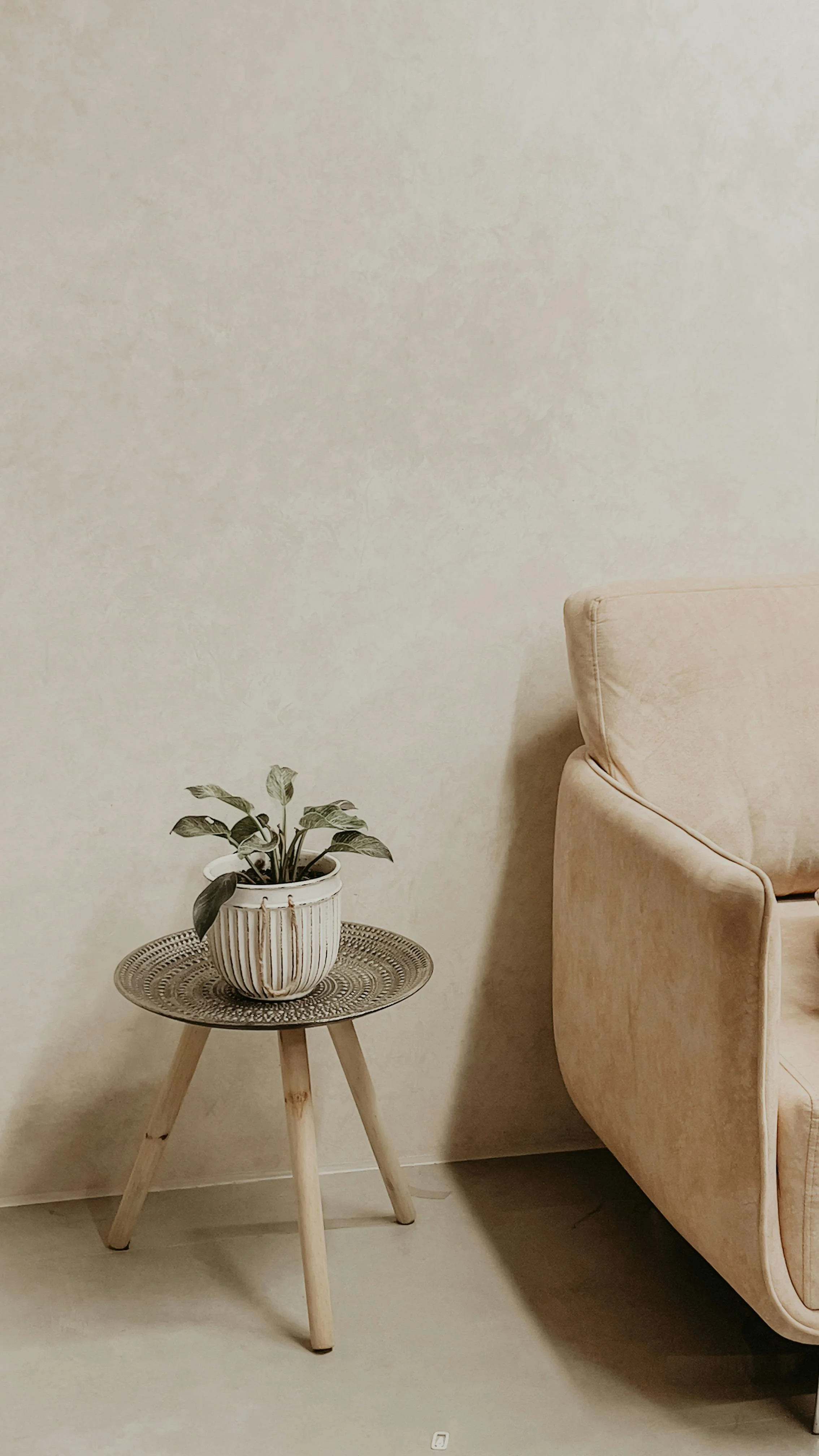 A small round wooden table with a woven top, holding a potted plant in a white ribbed ceramic pot, placed next to a cream-colored upholstered armchair with curved armrests, set against a plain off-white textured wall.