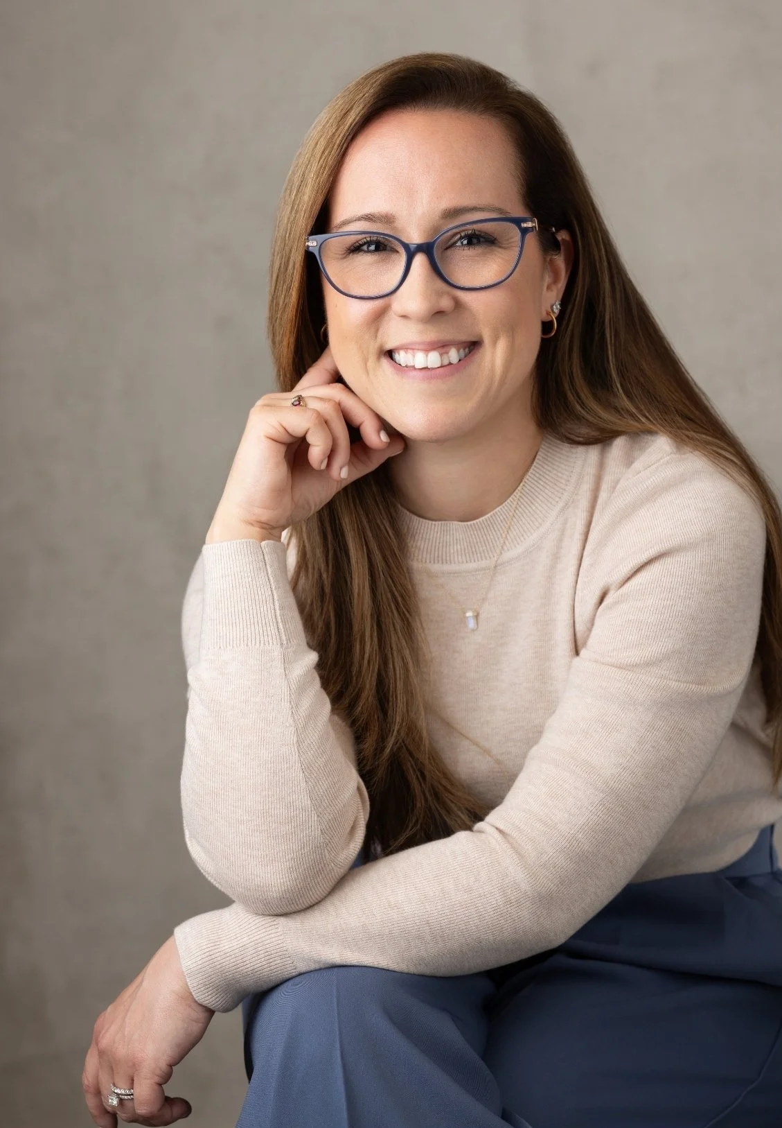 Dr. Kristina Burke, therapist based in Garden City, NY, with long brown hair, wearing glasses, a beige sweater, and blue pants, smiling and sitting with her head resting on her hand.