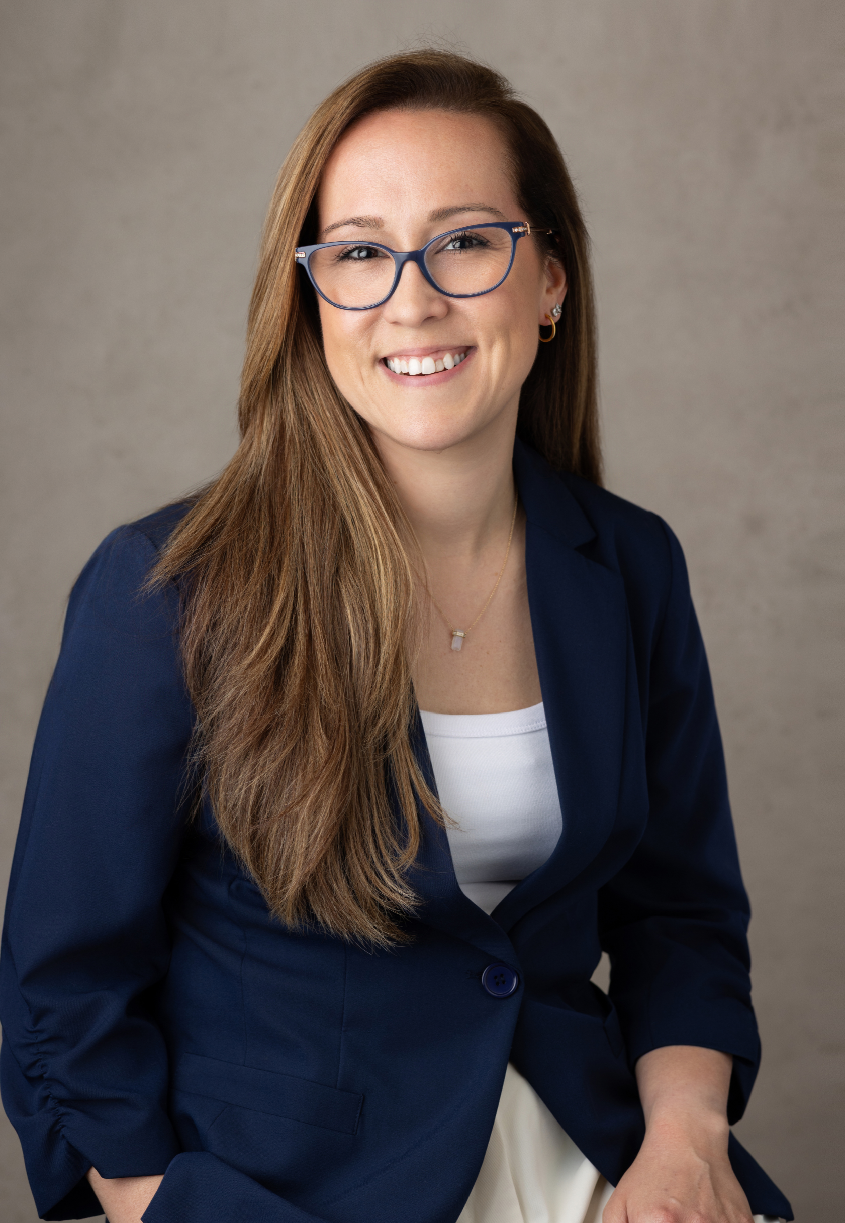 Dr. Kristina Burke, therapist based in Garden City, NY, with long brown hair and glasses smiling, wearing a navy blazer over a white top, seated against a gray background.