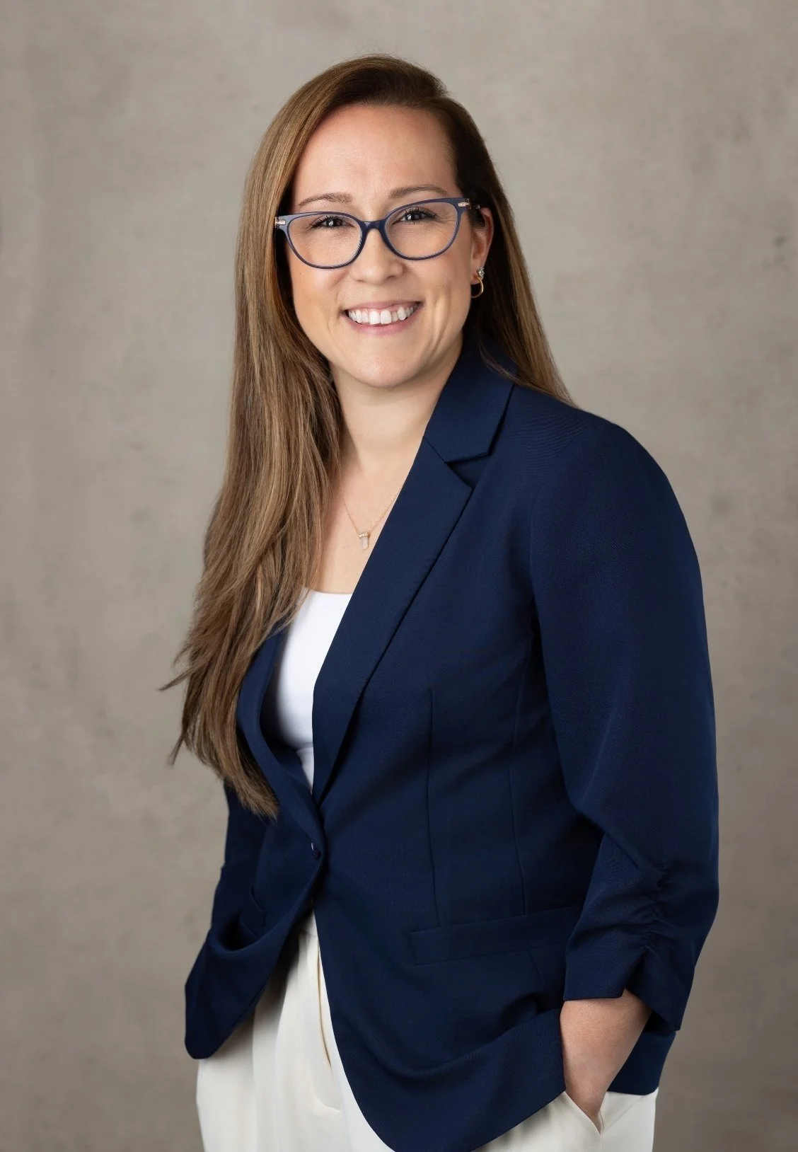 Dr. Kristina Burke, therapist based in Garden City, NY, with long brown hair, glasses, wearing a navy blazer over a white top, smiling with one hand in her pocket, against a plain gray background.