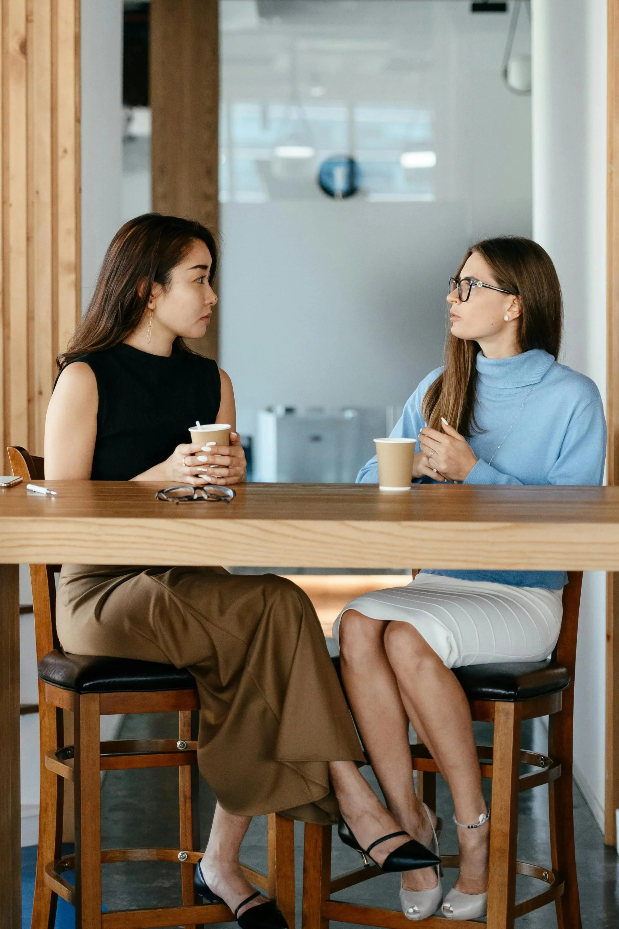 Two women are sitting at a high table in a modern café, facing each other. The woman on the left has long dark hair, tan skin, and is wearing a sleeveless black top and pants, holding a paper cup. The woman on the right has long brown hair, light skin, glasses, a light blue turtleneck sweater, and a beige skirt. They are engaged in conversation with cups on the table and glasses on the chairs.