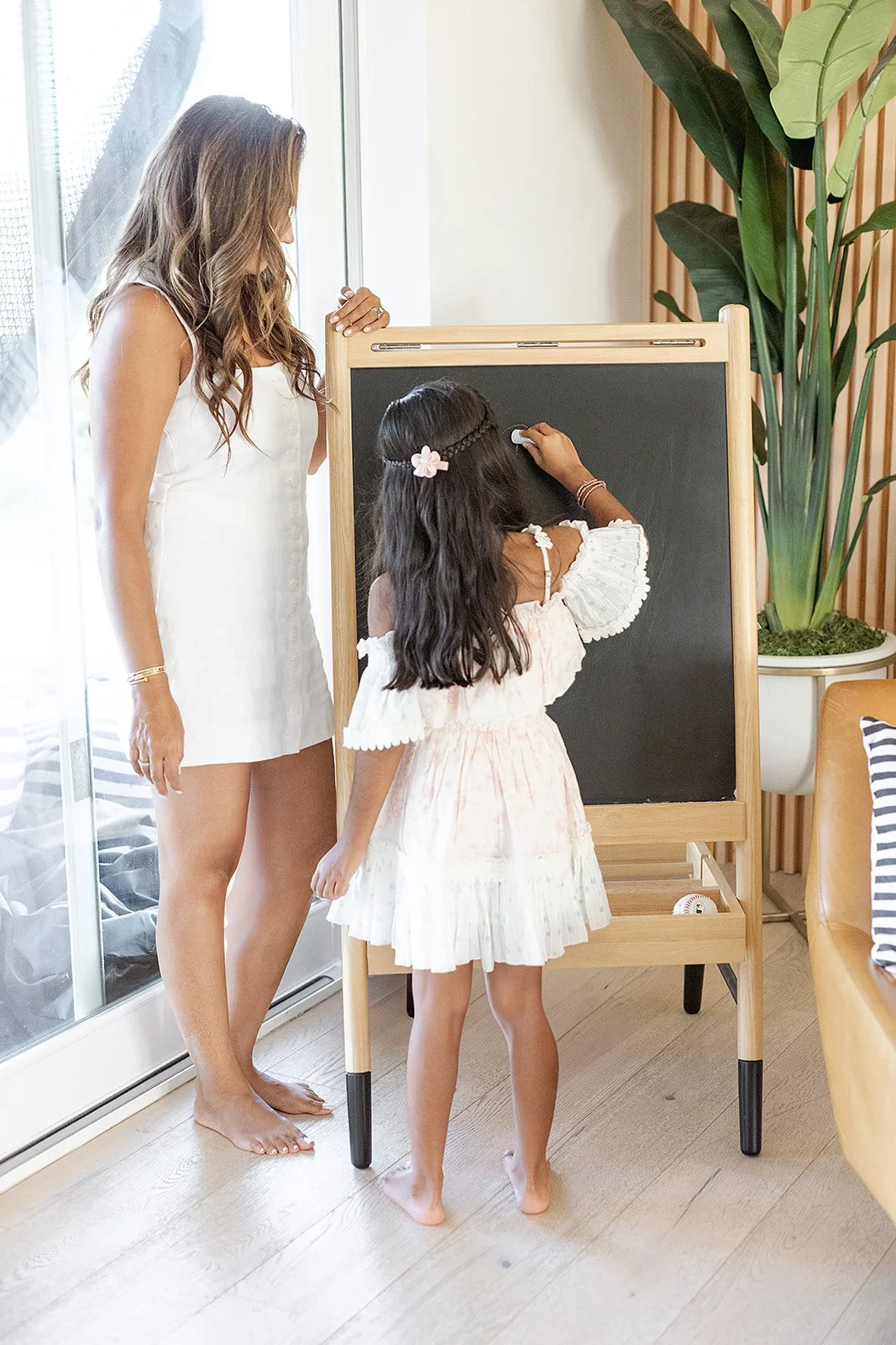 A woman and a young girl are standing near a chalkboard with the girl writing on it with chalk. The woman is looking on while the girl writes. There is a large potted plant nearby and a sliding glass door letting in natural light.