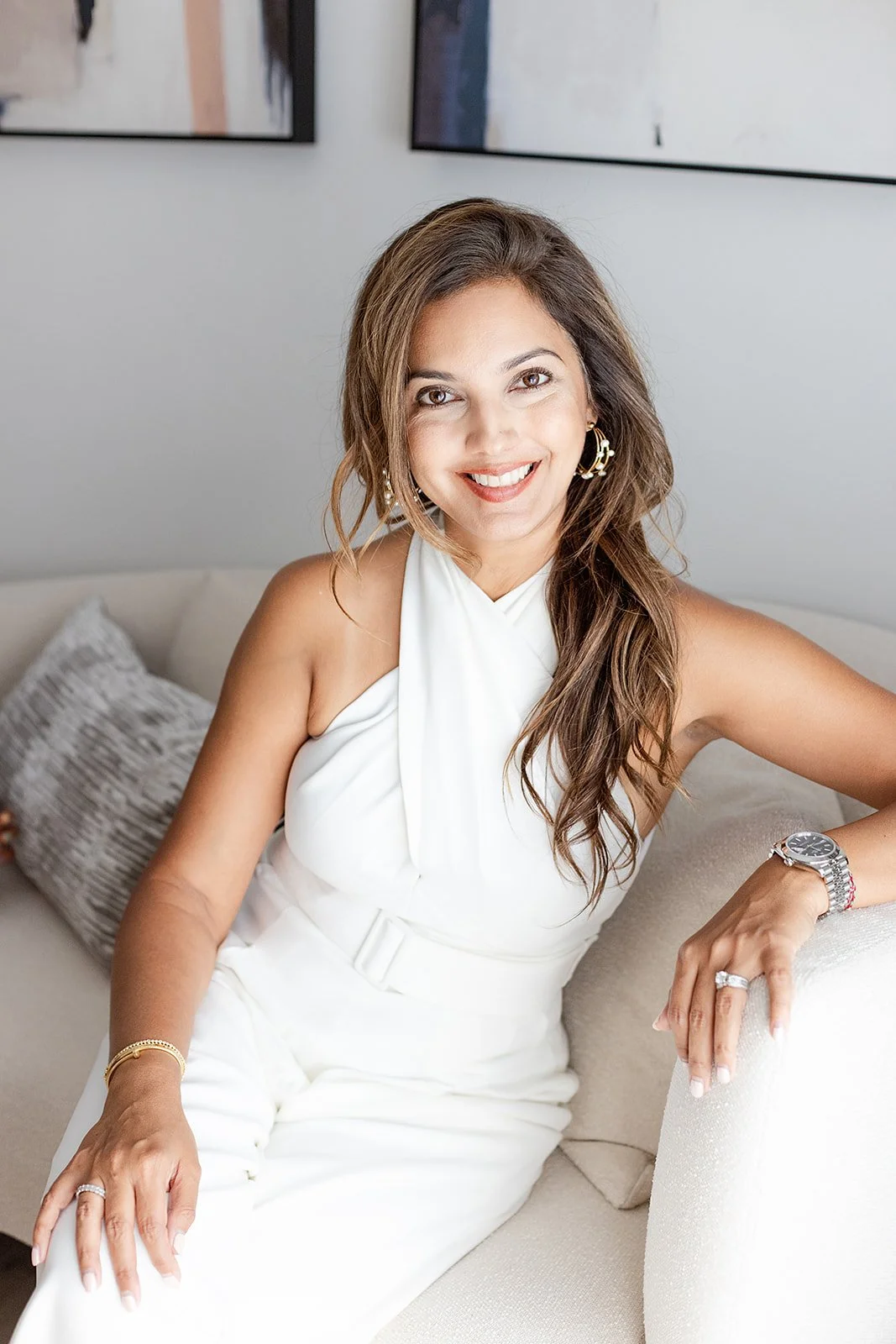 A woman with long brown hair in a white sleeveless dress, sitting on a beige couch, smiling at the camera.