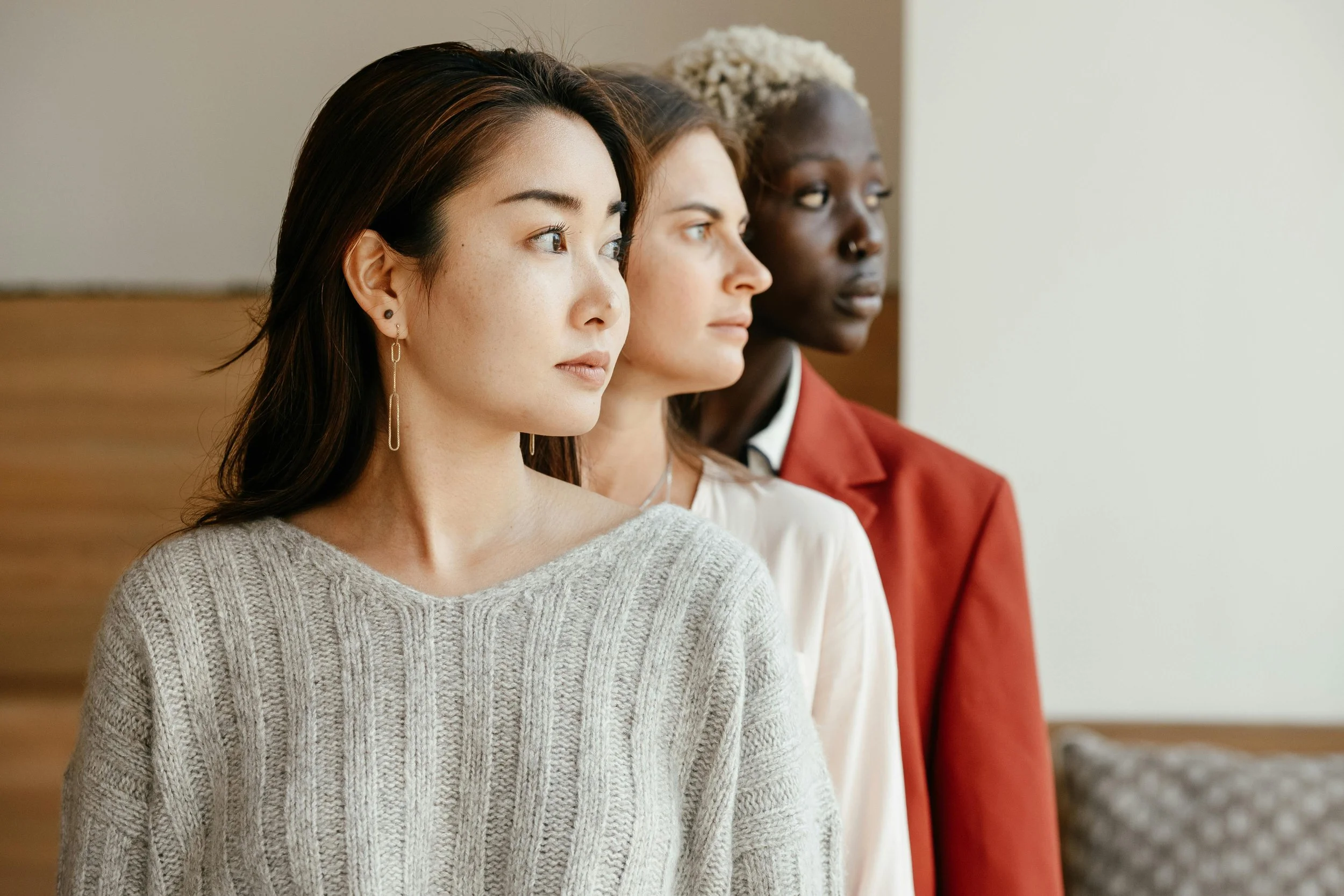 Three women stand side by side indoors, looking to the right. The woman in the foreground has long dark hair, light skin, and is wearing a gray knit sweater and earrings. The middle woman has curly dark hair, medium skin, and is wearing a white top. The woman in the background has short, platinum blonde hair, dark skin, and is wearing a red blazer.