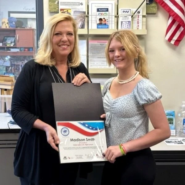 Two women, one older and one younger, smiling and holding a certificate of achievement, in an office or classroom setting with books and American flags in the background.