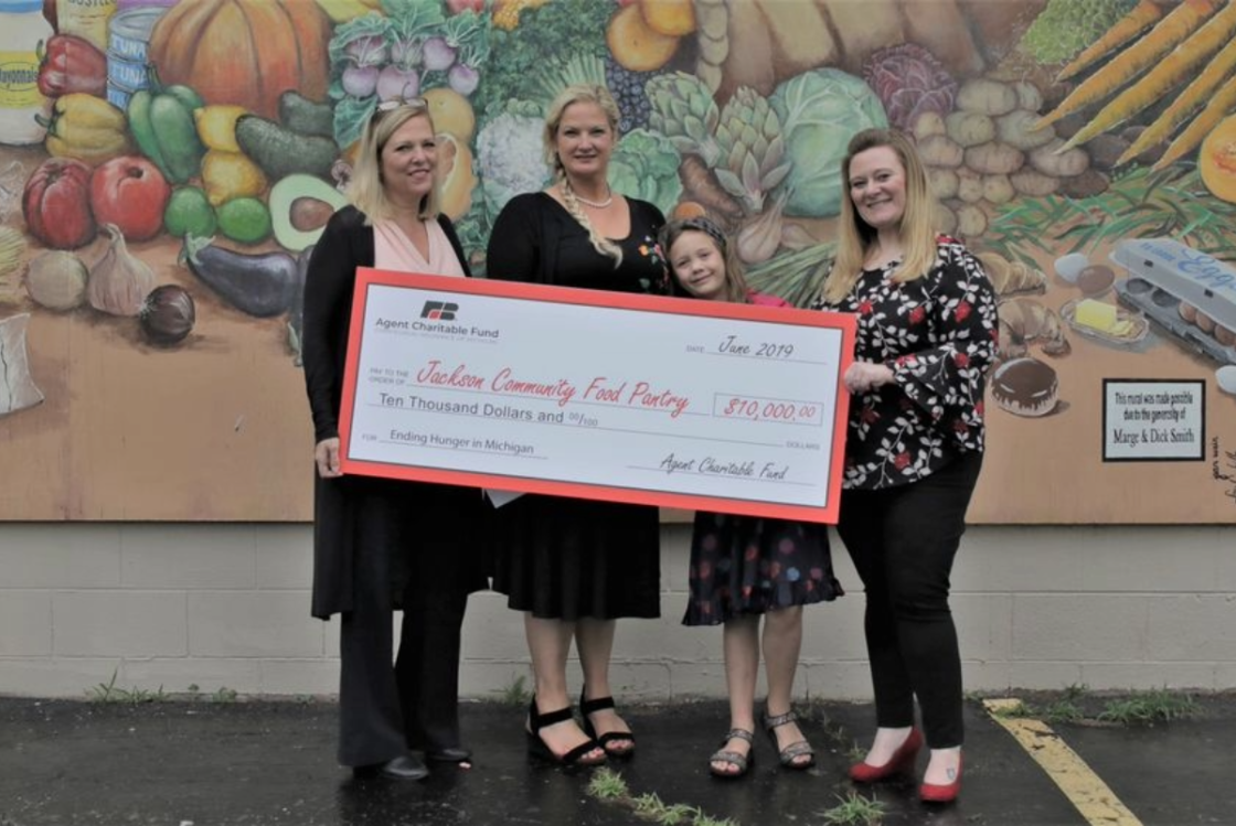 Four women and a girl holding a large check for $10,000 towards the Jackson Community Food Pantry. They are standing in front of a colorful mural of fruits and vegetables. The woman on the far right is wearing a black and white floral top, a girl in the center is wearing a pink top, and the woman on the far left is wearing a black jacket and pants.
