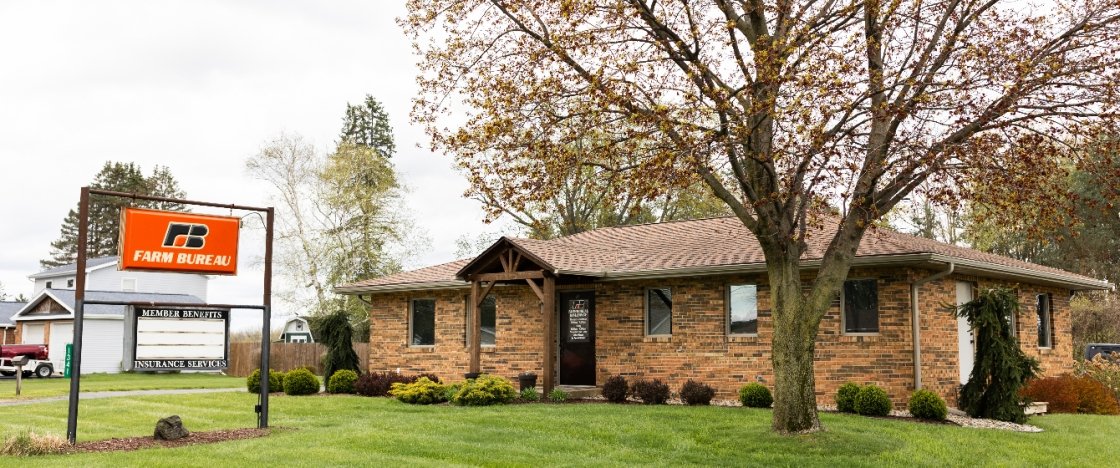 A brick building with a red-brown roof, surrounded by grass, trees, and bushes, with a sign for a farm bureau insurance agency in front.