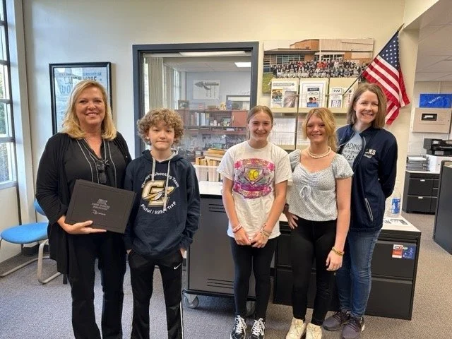 A group of five people standing together in an office or classroom with American flag and bookshelves behind them. The woman on the far left is holding a black folder or award. The group consists of one woman and four children or teenagers.