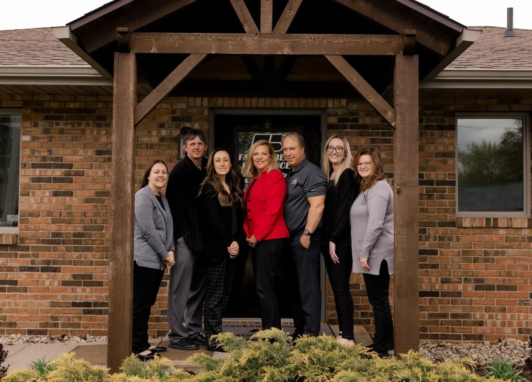 Group of seven people standing together outside a brick building under a wooden porch. The group includes men and women dressed in business casual attire, smiling and posing for the photo.