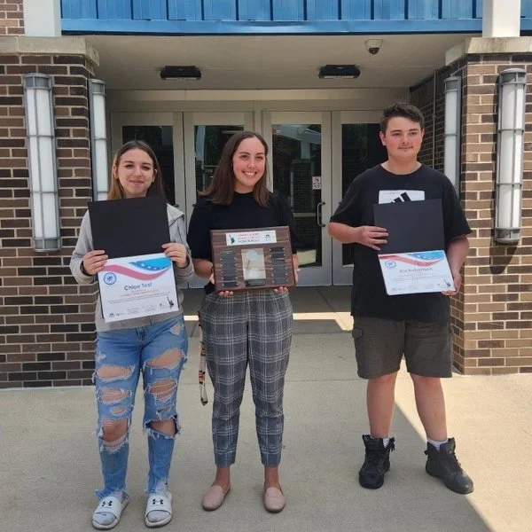 Three teenagers standing outside a building, each holding a certificate and a black folder. They are smiling, and there is a glass door behind them.