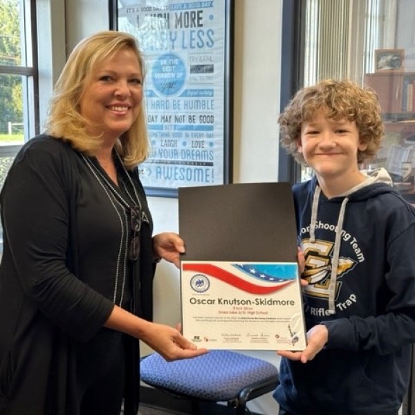 A woman and a young boy smiling and holding a certificate of achievement inside a room with large windows, blinds, and posters on the wall.