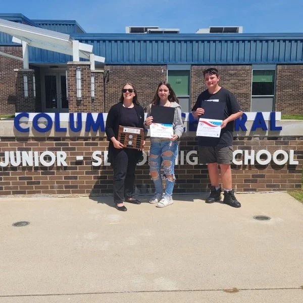 Three students standing in front of Columbia school sign, holding awards and certificates, on a sunny day.