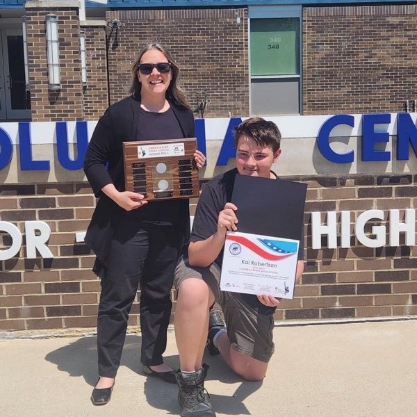 A woman and a boy standing and kneeling in front of a brick building. The woman is holding a plaque and the boy is holding a certificate, celebrating an achievement outside a volunteer or community center.