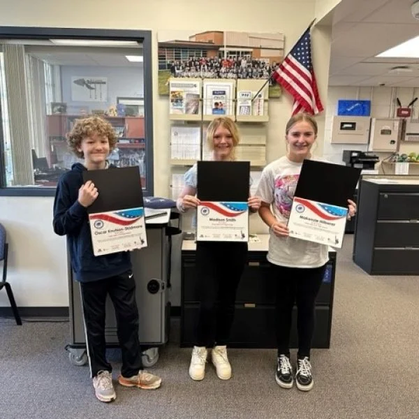 Three children holding certificates for the Odyssey of the Mind competition, standing indoors with an American flag behind them.