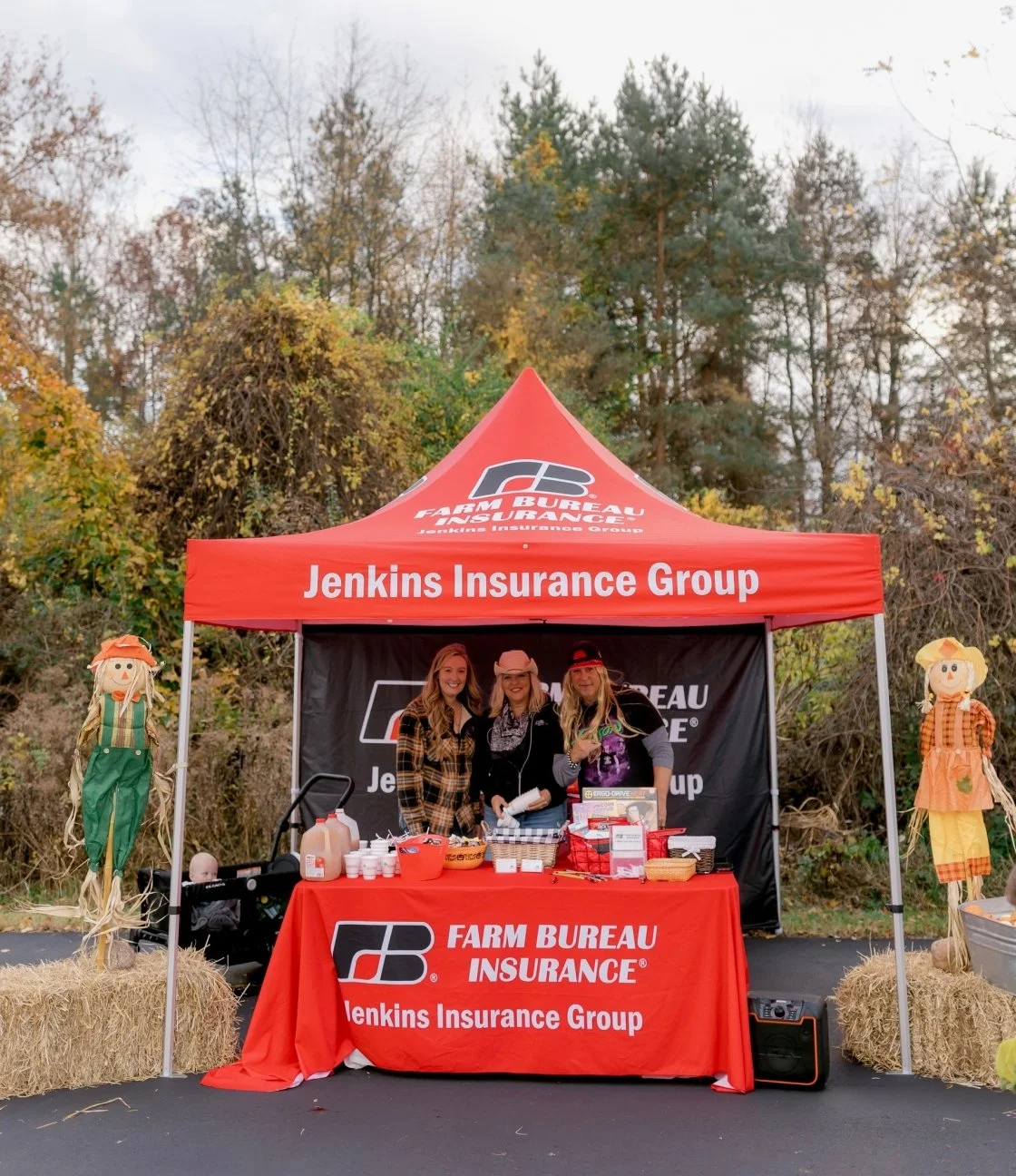 A red tent with 'Jenkins Insurance Group' and 'Farm Bureau Insurance' branding, set up outdoors with three women standing behind a table with promotional materials, surrounded by hay bales and scarecrows, with trees in the background.