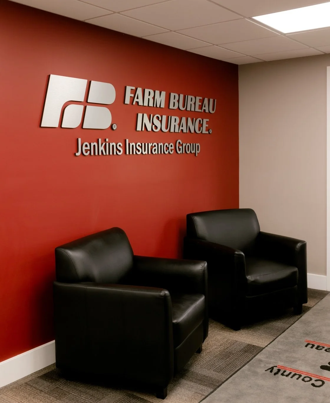 Lobby area with two black leather chairs against a red wall, featuring the Farm Bureau Insurance logo and the text 'Jenkins Insurance Group' in white letters.