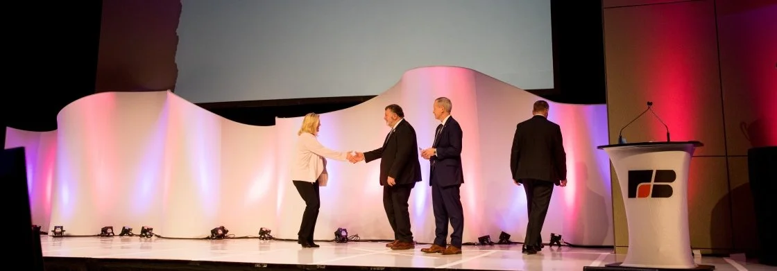 People on stage, with two women and two men, engaged in a handshake and conversation, in a conference or award ceremony setting.