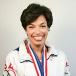 Close-up of smiling female athlete with short dark hair wearing medals and a white sports jacket, against a light blue background.