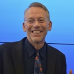 A smiling man wearing glasses and a dark blazer, standing in front of a blue background.