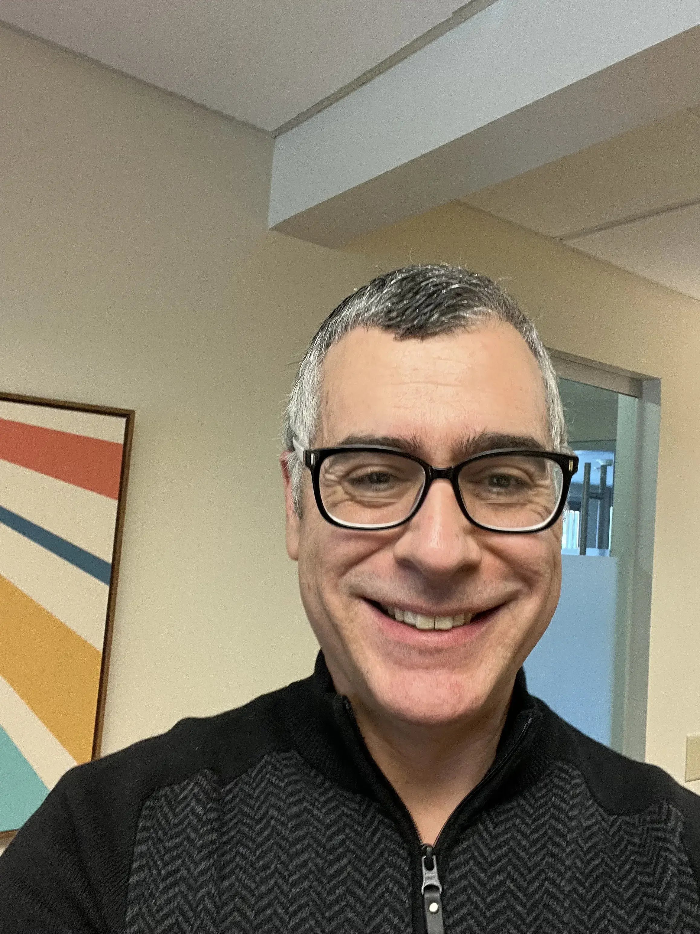 A smiling middle-aged man with gray hair and glasses wearing a black jacket, indoors with a colorful striped painting on the wall behind him.