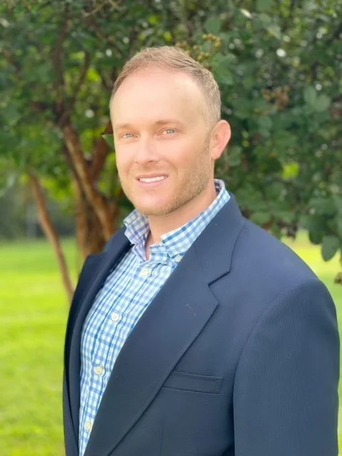 A man with short light hair and light skin, wearing a blue blazer and a blue checkered shirt, standing outdoors in front of a tree with green leaves.