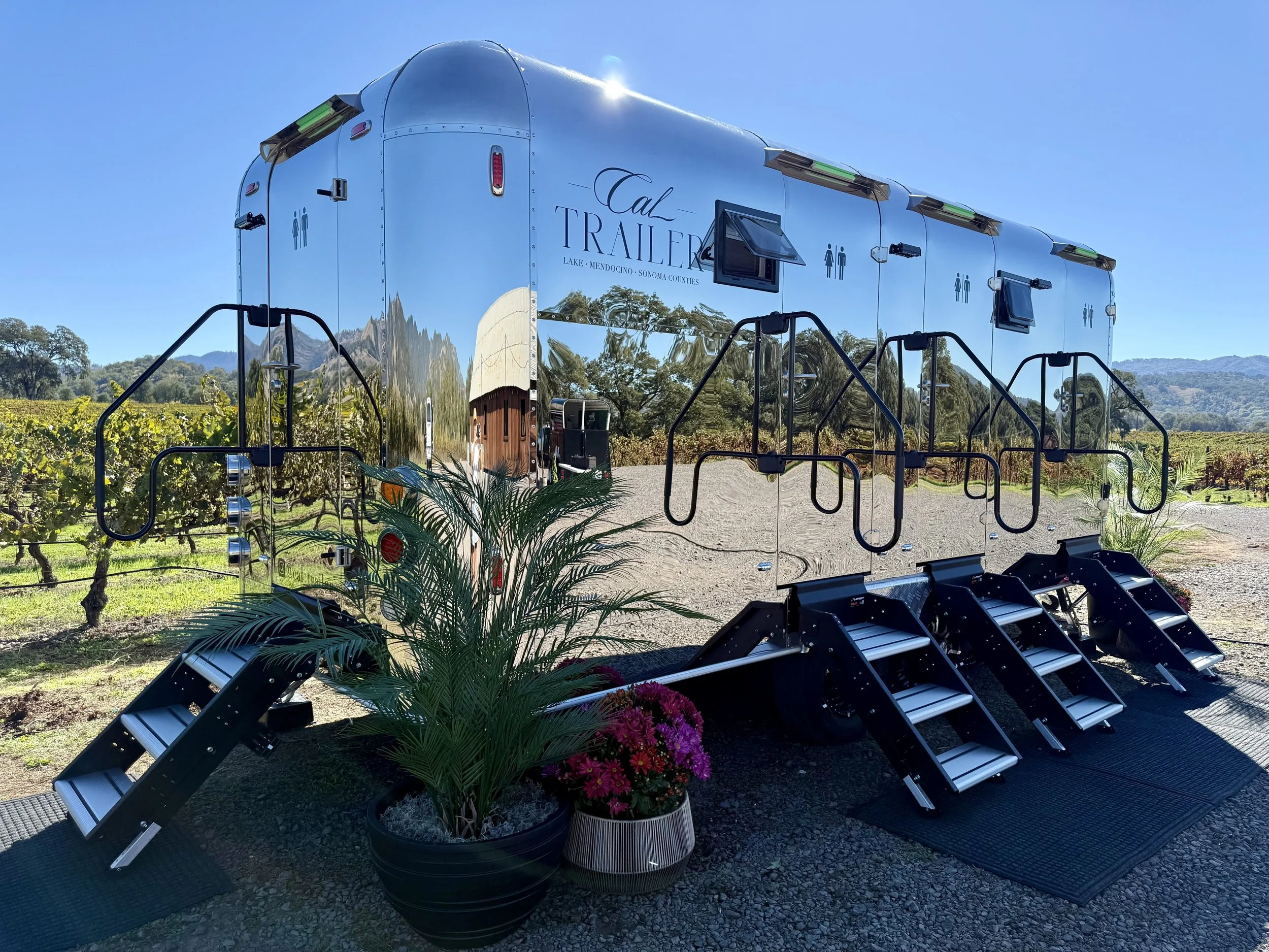 A shiny silver Airstream trailer with the words "Cal Trailer" and locations Lake, Mendocino, Sonoma Counties written on it. It is parked outdoors amidst trees, with outdoor stairs and flower pots in the foreground. The sky is clear and blue.